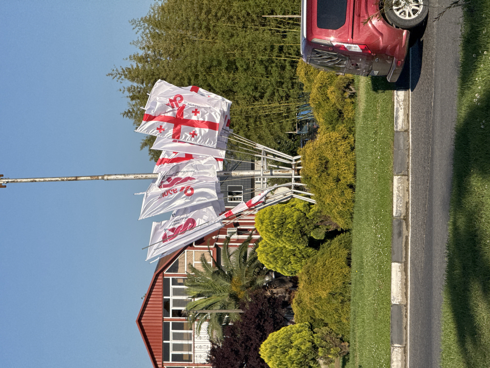 The image captures a scene in Batumi, Georgia, notable for its prominent display of Georgian national flags. Multiple white flags bearing the red St. George's cross are attached to a metal pole array. The backdrop includes a building with red roof and white-framed windows, a palm tree, various bushes and greenery, and bamboo stalks. A maroon-colored minivan is parked on the edge of a paved road with a grassy median. The sky is clear and blue.
