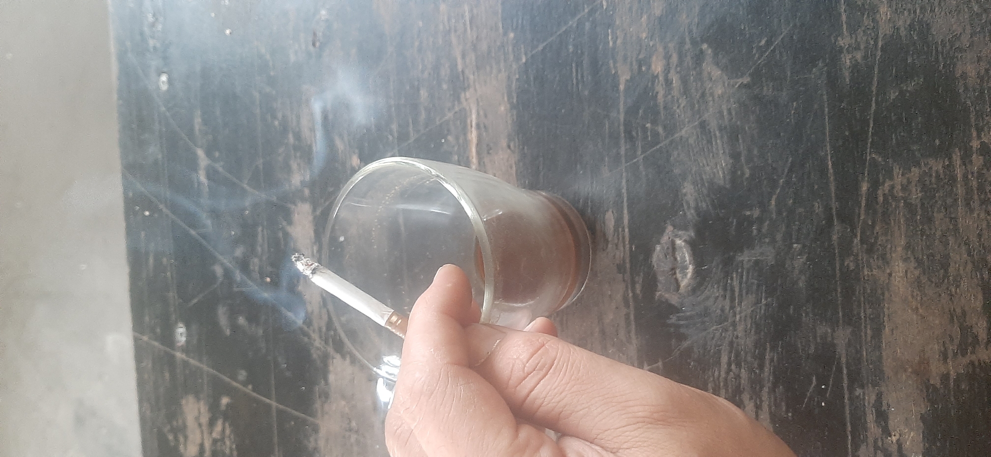 The image shows a close-up of a hand holding a lit cigarette over a glass cup of dark liquid, likely coffee or tea. The cigarette is actively burning and producing smoke, which is visible rising in the air. The hand is positioned on the left side of the frame, with the fingers wrapped around the stem of the cigarette and the thumb resting near the glass. The glass cup is transparent and contains a dark, opaque liquid that fills the bottom portion of the cup. The cup is resting on a dark, textured surface that appears to be made of wood, possibly a table with visible grain and wear marks. The background is out of focus, but suggests a muted, indoor or outdoor environment with soft lighting. There is no discernible urban landscape or specific geographical markers that would indicate a location within the city of Yessentukskaya, Russia. The scene is focused on the intimate action of smoking and drinking.