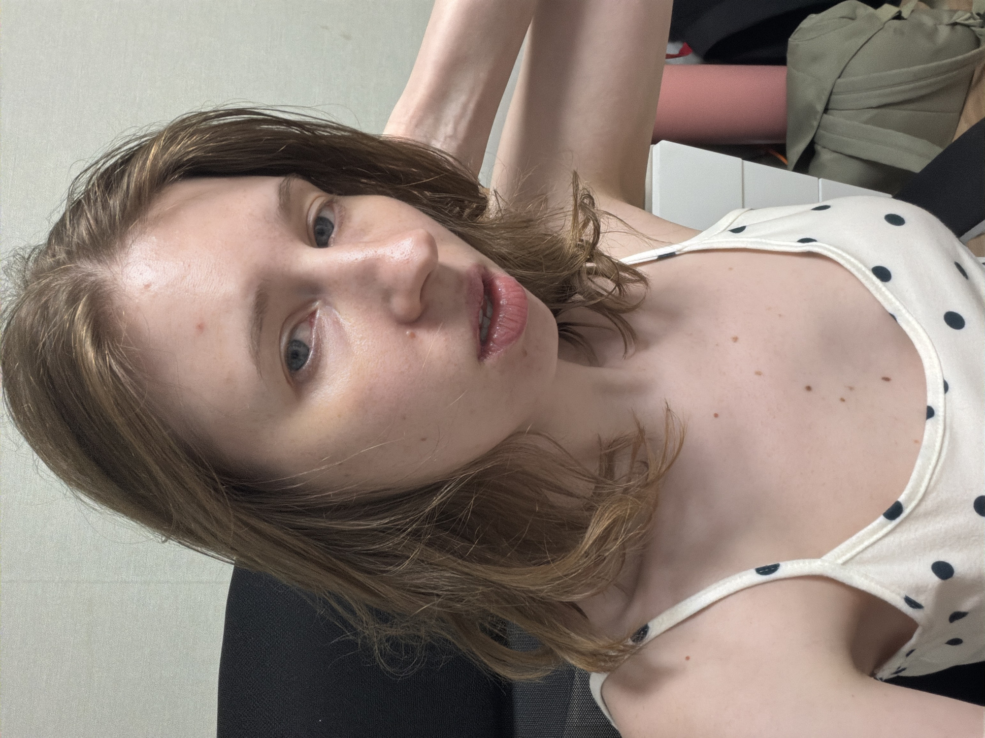 A young Caucasian woman with medium-length brown hair, wearing a white polka-dot camisole, is seated in a black office chair against a plain, light-colored wall. Her right hand is raised and resting on the back of her head. She is looking directly at the camera with a slightly parted mouth, and her skin appears shiny, possibly due to sweat or lotion. There are a few moles visible on her face and chest. In the background, a white set of drawers, a pink cylinder, and a folded green backpack are present. The lighting suggests an indoor setting, likely a home or office. No specific identifying features of Dongducheon-si are visible.
