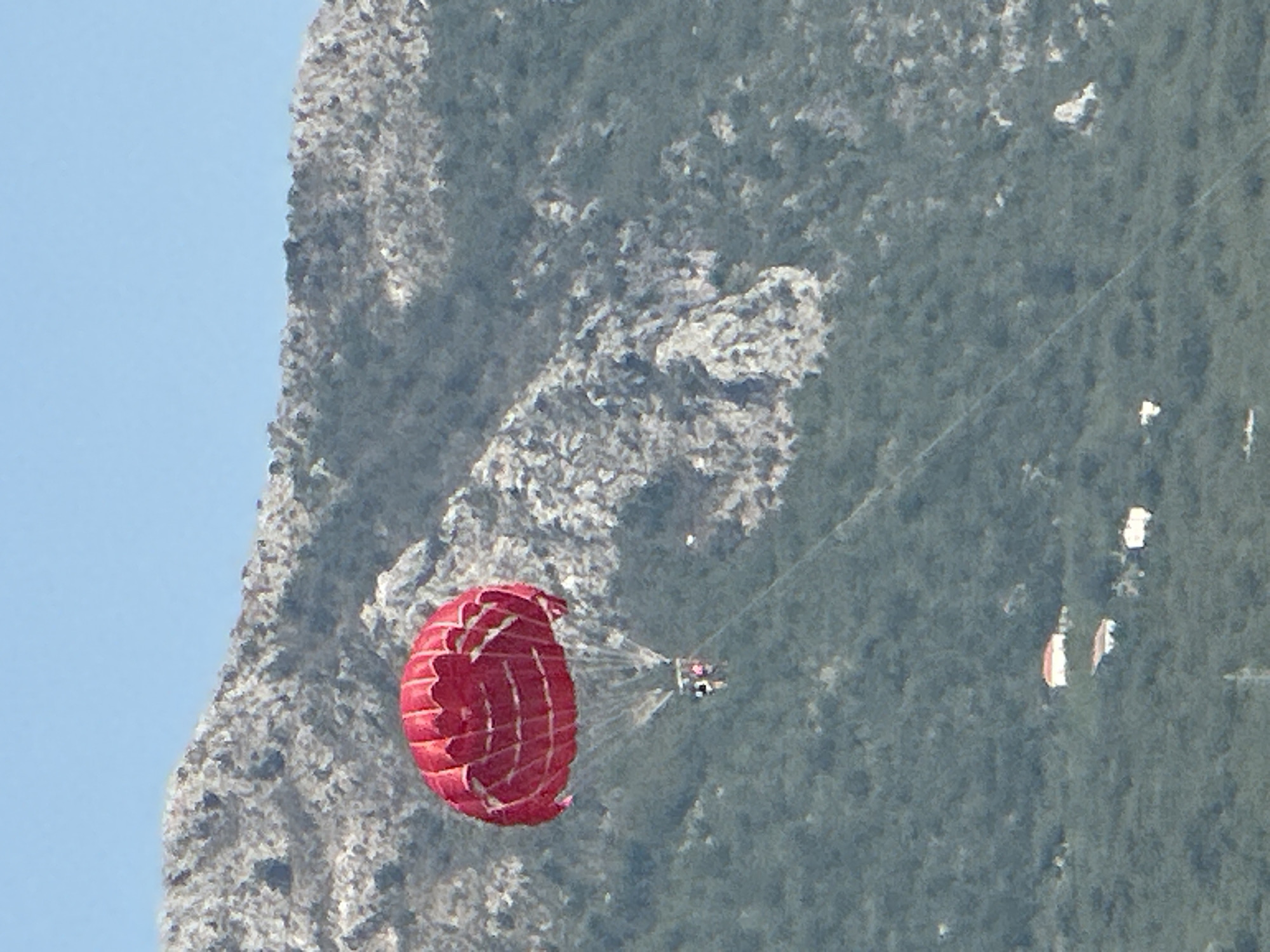 A red parachute with two people harnessed beneath it is suspended in the air against a backdrop of a rocky, tree-covered mountain and a clear blue sky. The parachute is predominantly red with white stitching creating a diamond pattern. The two individuals appear to be looking forward. Below them, the mountain slopes are covered with dense green foliage. Scattered across the lower portion of the mountain are a few small white buildings with red or white roofs. The overall scene suggests a recreational activity taking place in a mountainous coastal region, likely in or near Boreti, Montenegro, given the presence of the Mediterranean flora and architecture.