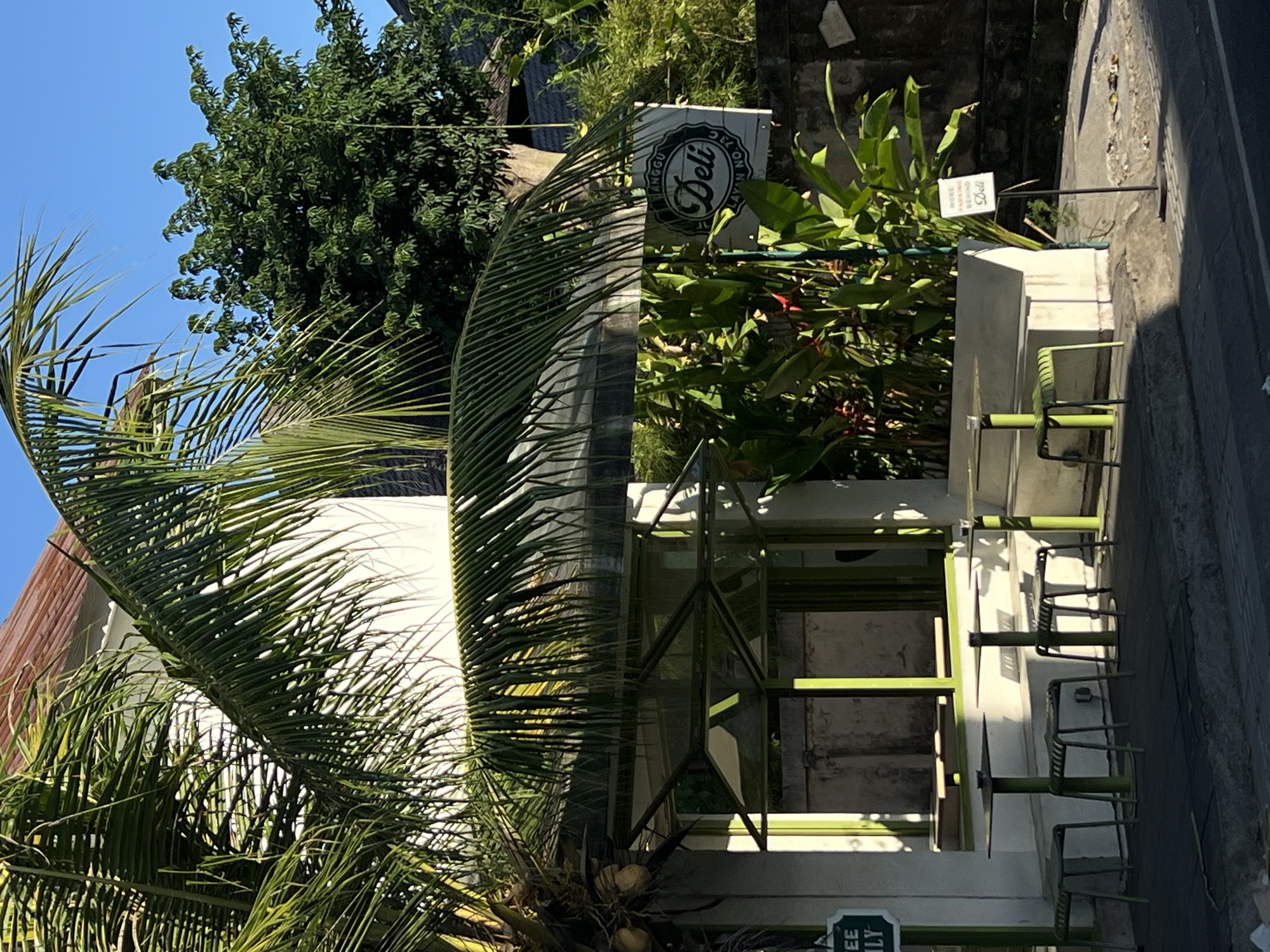 The image shows the exterior of a "Deli" establishment located in North Kuta, Indonesia. The building has a modern design with large windows and a green frame structure. Tables and chairs are set up outside, suggesting a cafe or restaurant setting. Lush tropical vegetation surrounds the building, including palm trees and various plants. A sign indicates "More Parking Behind Deli." The scene appears to be on a sunny day.