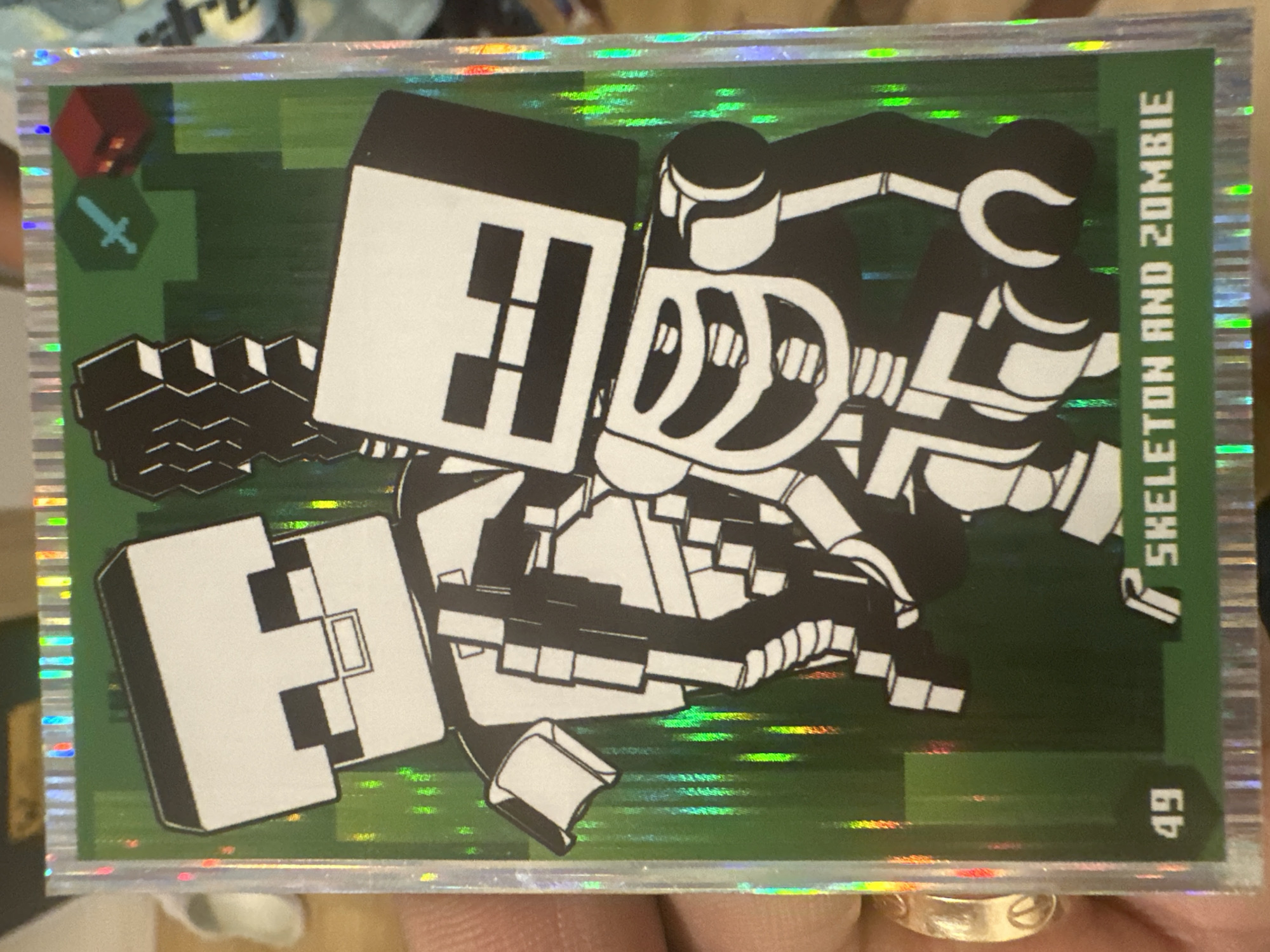 The image shows a collectible card featuring two characters from the video game Minecraft. The characters are a skeleton and a zombie, depicted in black and white pixelated art against a green, textured background that resembles grass blocks. The skeleton is on the left, with its ribcage and skull clearly visible, holding what appears to be a bone weapon. The zombie is on the right, also in a pixelated style, with its distinct blocky features. The card has a holographic border that reflects light. In the top right corner, there are small icons representing a sword and a red block, possibly indicating game elements. At the bottom left, the number "49" is displayed, and to the right of that, the text "SKELETON AND ZOMBIE" is written. The card is being held by a hand, with part of a ring visible on the ring finger. The background behind the card is blurred and appears to be an indoor setting. No location in Mainburg, Germany is depicted or suggested by the image.