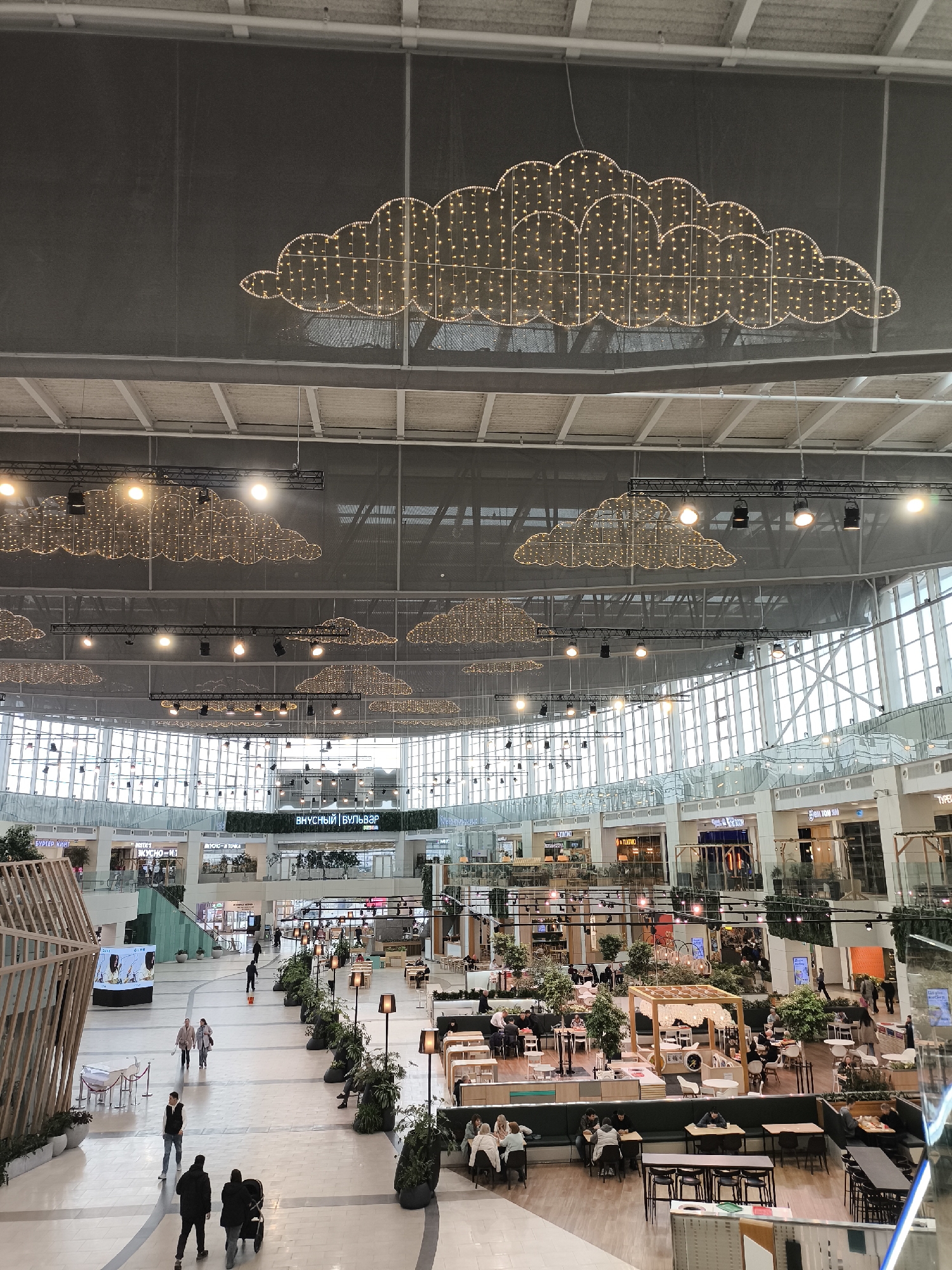 Here is a detailed description of the image:
The image captures a wide, interior view of a contemporary shopping mall. The location appears to be in Unknown, Unknown. Overhead, the ceiling is adorned with decorative light fixtures shaped like clouds. Natural light floods the scene through large windows situated along the mall's perimeter.
The interior is spacious, with a wide central walkway. Several small trees and other plants line the walkway, creating a park-like feel. Numerous people are visible, some strolling through the mall, while others are seated at tables in cafe-style setups.
The mall's architecture features multiple levels with stores and restaurants lining the perimeter. There are signs in an unknown language, indicating the names of establishments. Modern architectural elements are visible, including minimalist kiosks and wooden structures.