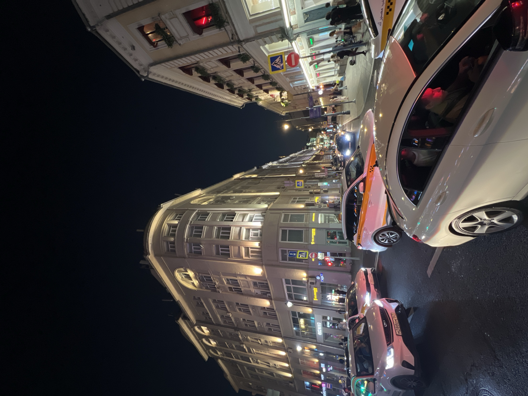 An eye-level, low-angle, full shot captures a street scene at night in Moscow, Russia, with traffic congestion and pedestrians. The foreground features the back of a white Mercedes-Benz sedan, its tinted windows reflecting the surrounding lights. To its left, a white Geely Coolray SUV, with license plate M288EM 147, is stopped in traffic. Behind it, a white taxi with orange stripes is also stationary. Further down the street, a dark blue car with its headlights on is visible.
The buildings flanking the street are illuminated, casting warm light onto the facades. The building on the left is a large, cream-colored structure with multiple floors and arched windows. The building on the right is a lighter cream color, with red awnings above some of its windows and decorative elements along its roofline. Pedestrians are scattered along the sidewalks, some walking and others gathered in small groups. Traffic lights and street signs are visible, indicating a busy urban intersection. The overall impression is one of a vibrant city at night, with the flow of traffic and pedestrian activity creating a dynamic atmosphere.