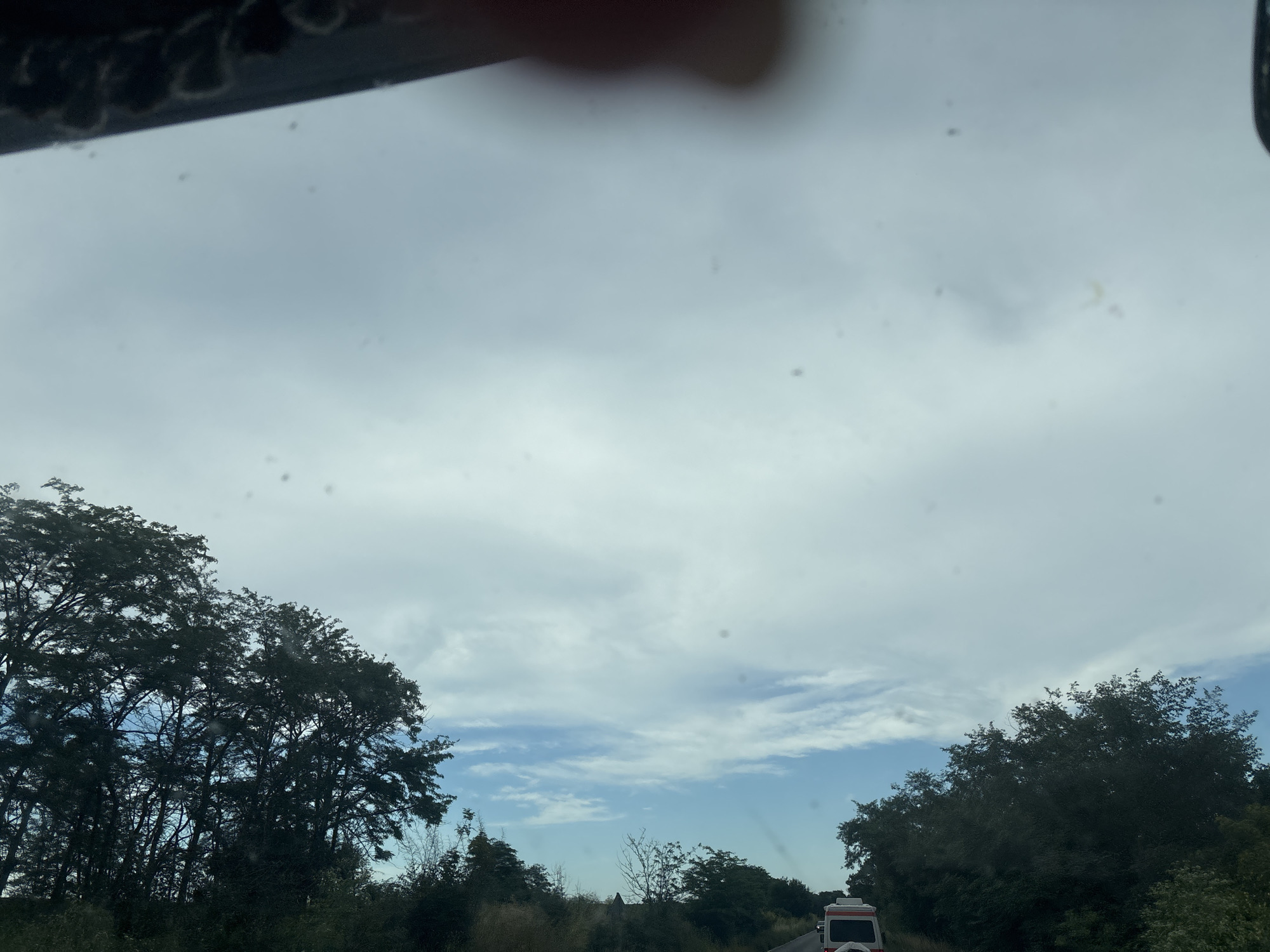 The image depicts a view from inside a vehicle, looking out at a cloudy sky over a landscape of trees and a road. On the left side of the image, a dense cluster of dark green trees is visible. In the distance, a light-colored vehicle, possibly a van or camper, is seen on the right side of the road. The road itself is paved and appears to be in a rural or suburban area, with vegetation lining its sides. The sky is predominantly filled with thick, gray clouds, though patches of blue sky with lighter clouds can be seen, particularly towards the horizon. The overall impression is that of a somewhat overcast day, possibly in the late afternoon or early morning, given the lighting. The scene is likely captured while driving, as suggested by the perspective and the blurry details of the sky and surroundings. The image has a number of small dark spots, possibly dust or water droplets on the windshield. There is no discernible city infrastructure visible. The image may have been taken in the vicinity of Cherni Vrah, Bulgaria, but this cannot be confirmed from the visual evidence alone.
