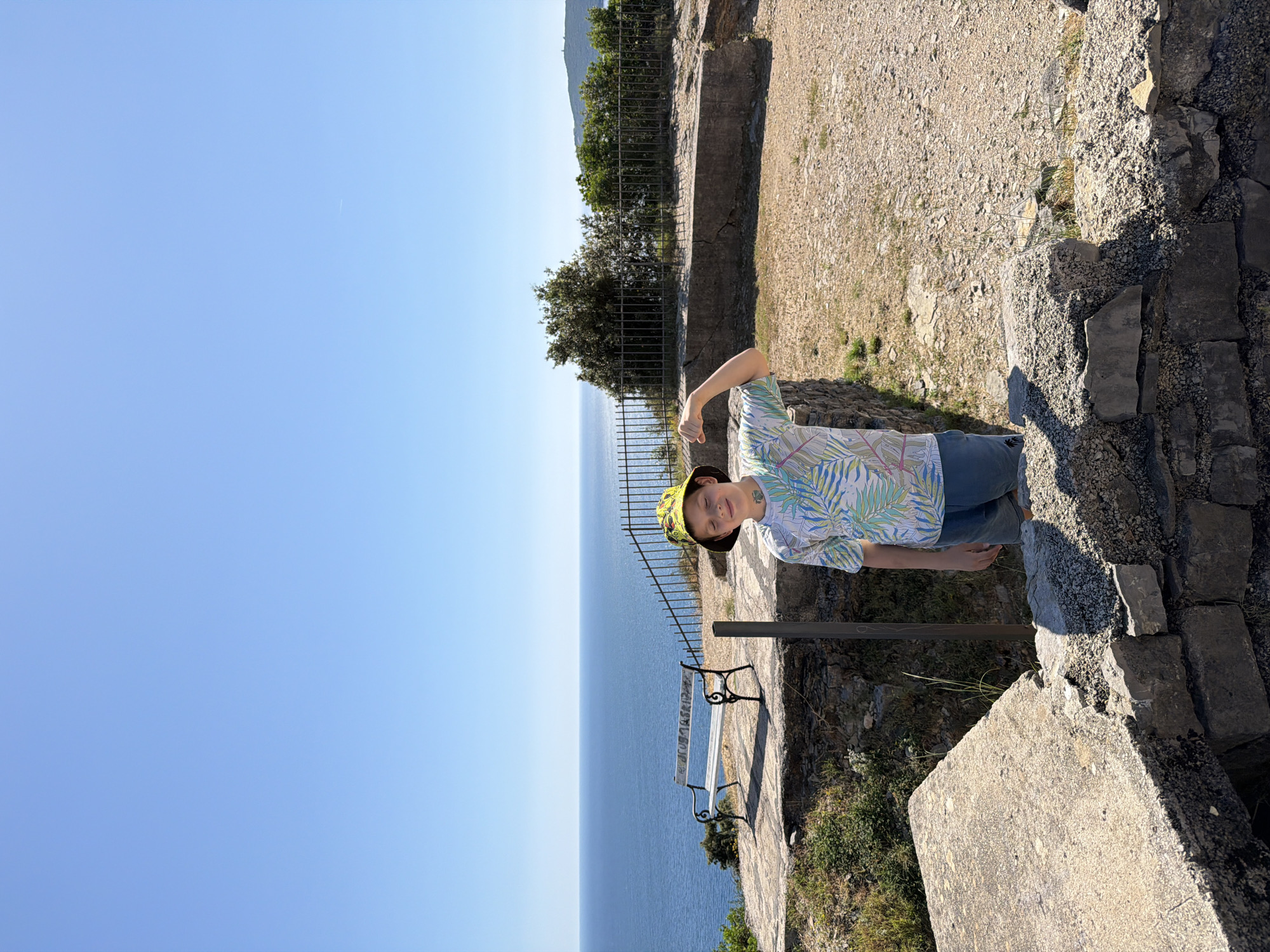 The image depicts an elevated coastal viewpoint under a clear, cloudless blue sky, consistent with a sunny day in Budva, Montenegro.
In the foreground, a single light-skinned young male individual, appearing to be a child or pre-teen, stands slightly right of center, angled towards the viewer. He wears a yellow and green patterned bucket hat, a short-sleeved t-shirt with a tropical leaf motif (comprising green, blue, white, and pinkish tones), and blue denim shorts. His right arm is bent at the elbow, with the hand fisted and held upwards in a flexing posture. His eyes are closed or squinted, and a subtle smile is present. A small, dark mark, resembling a temporary tattoo or drawing, is visible on his neck below his right ear. The immediate ground level in the foreground consists of rugged stone and concrete structures, appearing weathered.
Behind the individual, the midground features a white metal park bench with dark ornate supports, positioned on a concrete slab that overlooks the sea. The backrest of the bench displays visible graffiti. A dark metal fence extends across the scene, delineating the edge of the elevated platform from the descent to the water. Sparse vegetation, including bushes and small trees, grows behind the fence, particularly prominent on the right side.
The background is dominated by a vast expanse of calm, deep blue sea meeting a clear, bright blue horizon. On the distant right, a green landmass or island is visible, featuring a small white structure, possibly a lighthouse or communication tower, at its apex. The scene is illuminated by bright sunlight, casting distinct shadows. The combination of a fortified coastal outlook, the expansive sea view, and distant landforms aligns with locations found in Budva, Montenegro.