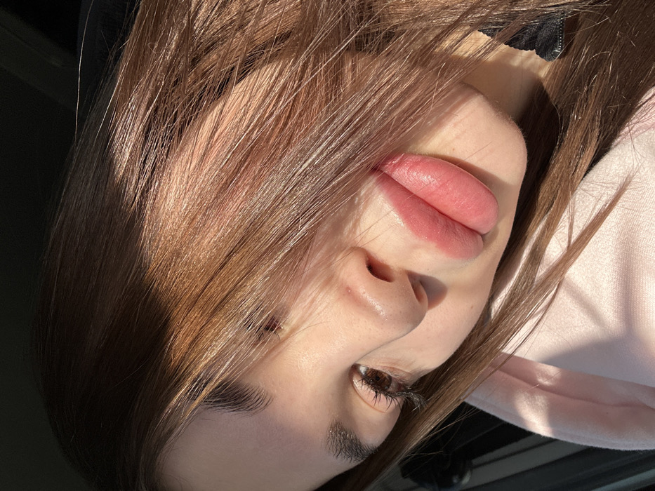The image presents a close-up, angled portrait of a single individual, likely a female, from the upper chest upwards. The subject has medium brown hair, with strands falling across the upper right portion of her face (viewer's left). Direct sunlight illuminates the upper left quadrant of the image, including portions of her hair and the right side of her face, creating a strong contrast with shadowed areas. Her left eye (viewer's right) is visible, featuring dark eyelashes and a light brown iris. The bridge and tip of her nose are visible. Her lips are full, exhibiting a pink hue, and appear slightly parted. Her skin is fair. She is wearing a light pink garment, possibly a hoodie or sweatshirt, visible on her lower left. A dark, indistinct fabric is visible near her neck on the lower right. The background is dark and out of focus, providing no specific environmental details. The location of Votkinsk, Russia, cannot be determined from the visual information within this image.