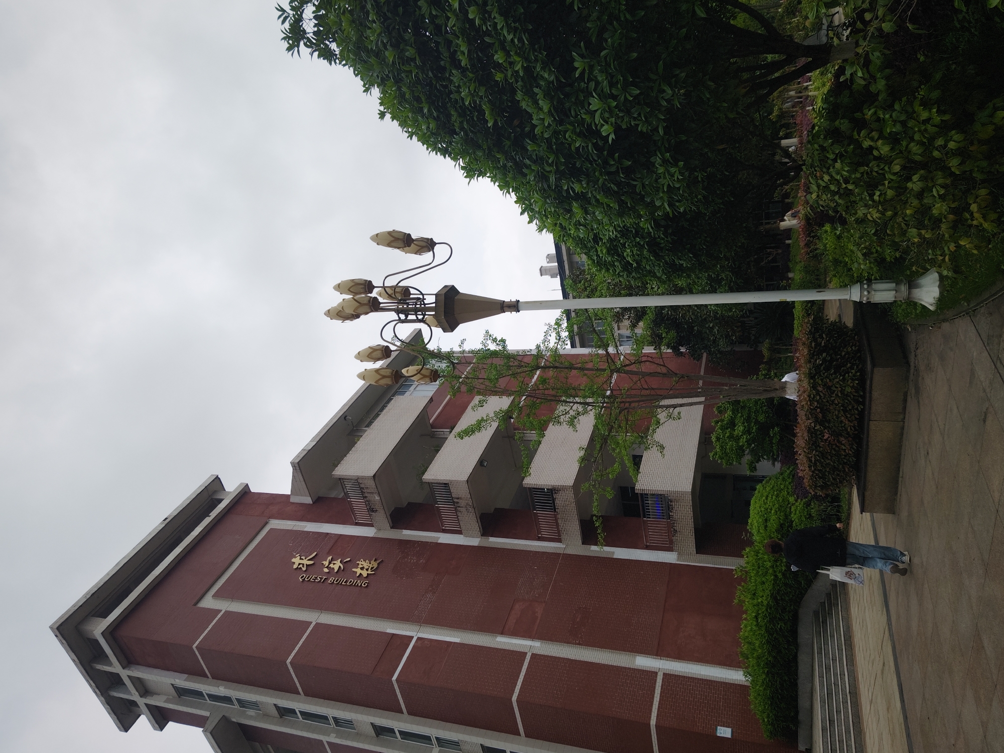 The image depicts the exterior of the Quest Building, located in the Chongqing High-tech Zone, China. The building is a brick structure with multiple stories and balconies. A lamp post stands in front of the building. A person is walking on the sidewalk near the entrance, beside the green bushes. The sky is overcast, and lush greenery is present in the background.