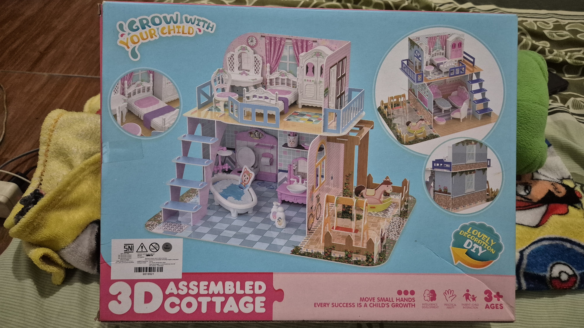 The image shows the box of a 3D assembled cottage toy, likely a dollhouse. The box is light blue with pink accents and features a detailed illustration of the assembled toy, which depicts a multi-story cottage with furniture and accessories like a bed, dresser, bathtub, and rocking horse. Smaller circular insets on the box show different configurations or views of the toy. The text on the box includes "GROW WITH YOUR CHILD," "ASSEMBLED COTTAGE," "LOVELY DECORATION DIY," and icons indicating benefits like "INTELLIGENCE DEVELOPMENT," "PRACTICAL ABILITY," "PARENT-CHILD INTERACTION," and "3+ AGES." A safety certification mark, "SNI," is visible in the lower left corner. The box is lying flat on a surface covered with a patterned fabric, possibly a blanket or bedsheet. In the background, to the left, a yellow towel or blanket is partially visible, along with some white electronic cables. The lighting suggests an indoor environment. The question asks for the location in Karawang, Indonesia, but no visual cues in the image provide any information about its specific geographical location, including within Karawang, Indonesia.