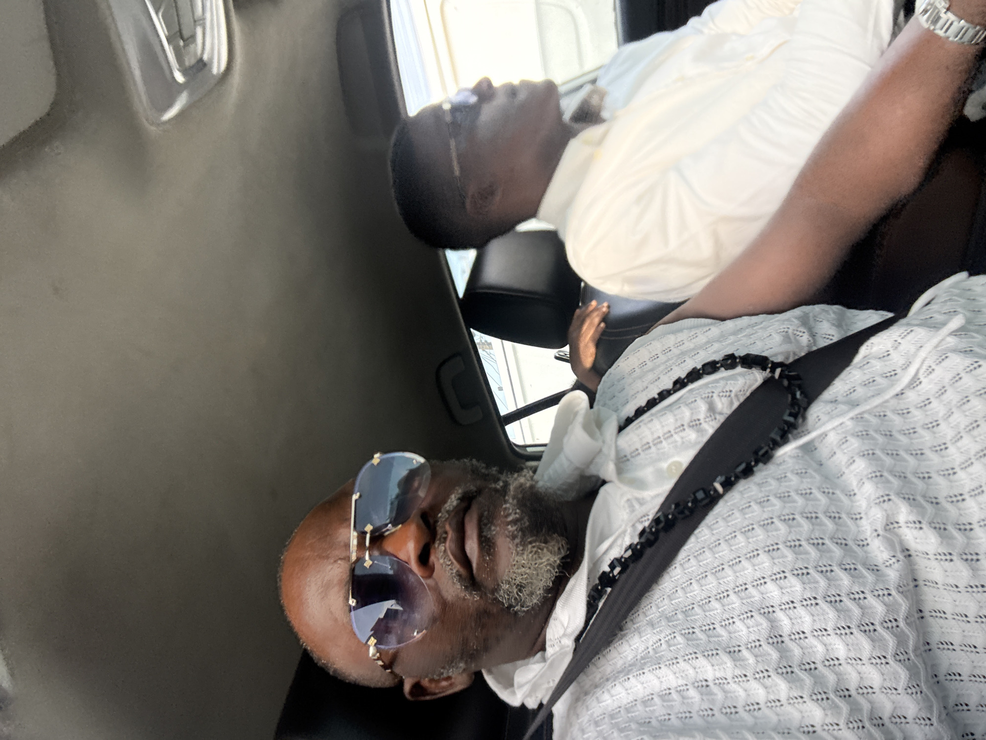 The image shows two Black men inside a vehicle. One man is wearing large sunglasses, a white patterned shirt, and a seatbelt. The other man is wearing a white shirt and glasses. The location is potentially Ado, Nigeria, based on the request, but this cannot be confirmed from the image itself. The scene appears to be inside a car, with interior features visible. The men are passengers, likely riding in the vehicle.