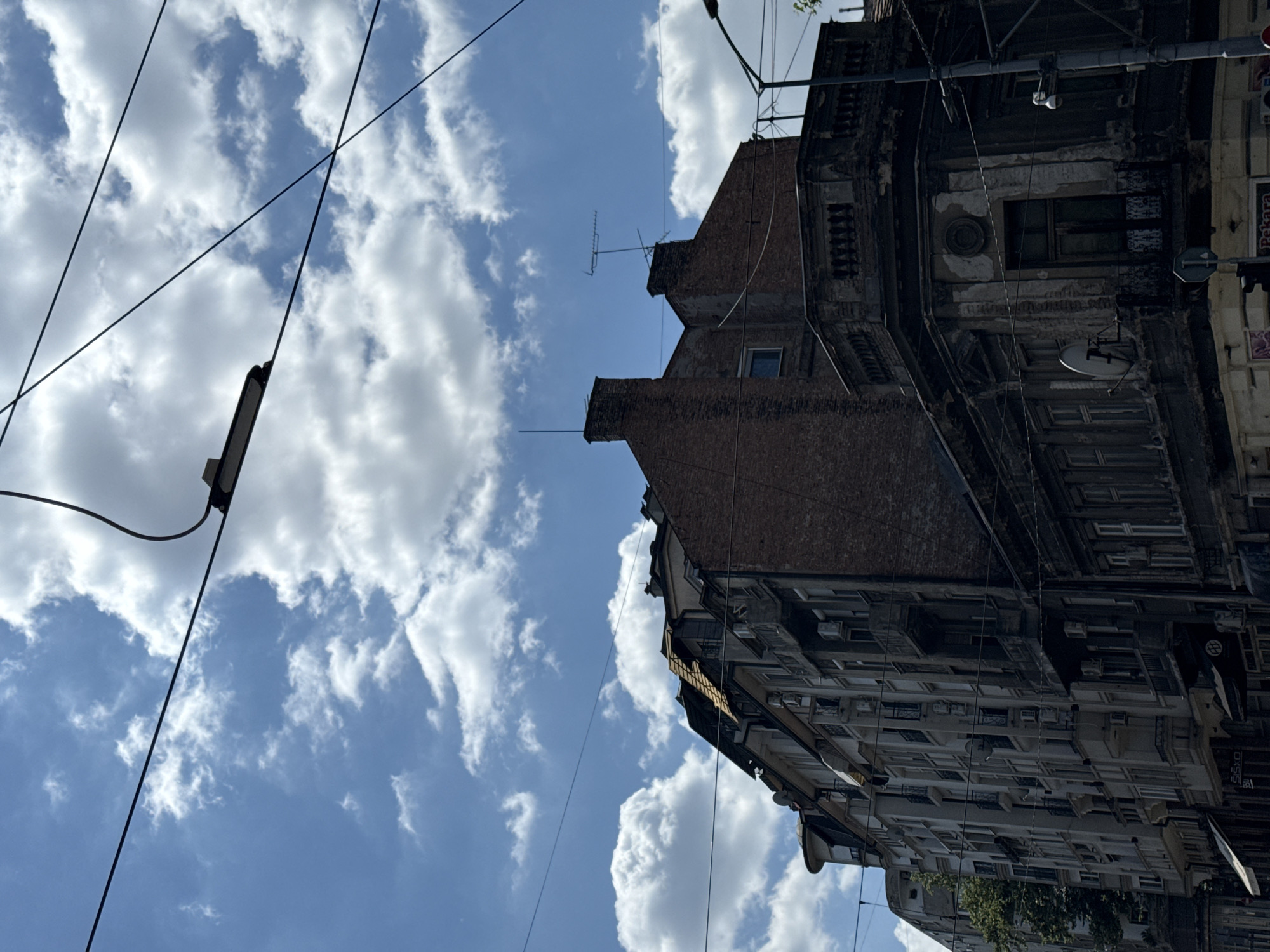 The image is a low-angle shot of urban architecture against a cloudy sky, taken in Belgrade, Serbia. The perspective is such that the buildings appear to be tilting upwards, and the sky occupies a significant portion of the frame.
The main subjects are two distinct buildings. The foreground building is older and appears to be in a state of disrepair, with crumbling brickwork, visible plaster damage, and weathered facades. It features multiple windows, some with visible shutters or ornate frames, and an antenna atop one of its brick chimneys. There are also several electrical wires and what appears to be a street lamp hanging in the sky.
Behind and to the right of this older building is a taller, more modern building with a repetitive facade of numerous windows, balconies, and air conditioning units. This building shows more signs of ongoing habitation, though some areas also appear aged.
The sky is a mix of bright blue and large, white cumulus clouds, suggesting a partly cloudy day. The lighting indicates it's daytime, likely late morning or early afternoon, given the brightness.
The overall mood is somewhat stark, focusing on the juxtaposition of old and new urban development. There are no people visible. The activity taking place is simply the existence of the urban environment.
Some very faint text is visible on signage on the modern building to the right, but it is too blurry to be deciphered. The presence of the antenna and electrical infrastructure are common urban features. The architectural style of the older building suggests a historical European city, consistent with the Belgrade context.