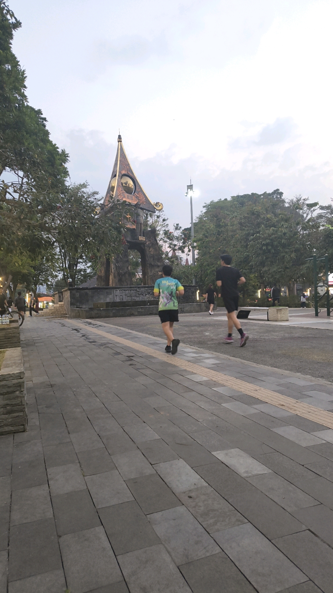 The image depicts an outdoor scene in Salatiga, Indonesia, likely in a public park or plaza. In the foreground, a paved walkway made of gray square and rectangular tiles dominates the lower half of the frame. A strip of yellow tactile paving runs parallel to the walkway. Two individuals are jogging on the walkway, moving away from the camera. The person on the left is wearing a multi-colored t-shirt and black shorts, while the person on the right is wearing a black t-shirt and black shorts.
In the background, a large, ornate monument with a distinctive, pointed roof structure stands prominently. This monument appears to be made of dark stone and metal, with decorative elements. Trees with lush green foliage surround the monument and line the sides of the walkway, indicating a park-like setting. Several streetlights and other decorative lights are visible, some illuminating the area. Other people can be seen in the distance, engaged in various activities, suggesting a public gathering space. The sky is overcast and appears to be late afternoon or early evening. The overall impression is one of a public space being utilized for recreation and leisure.