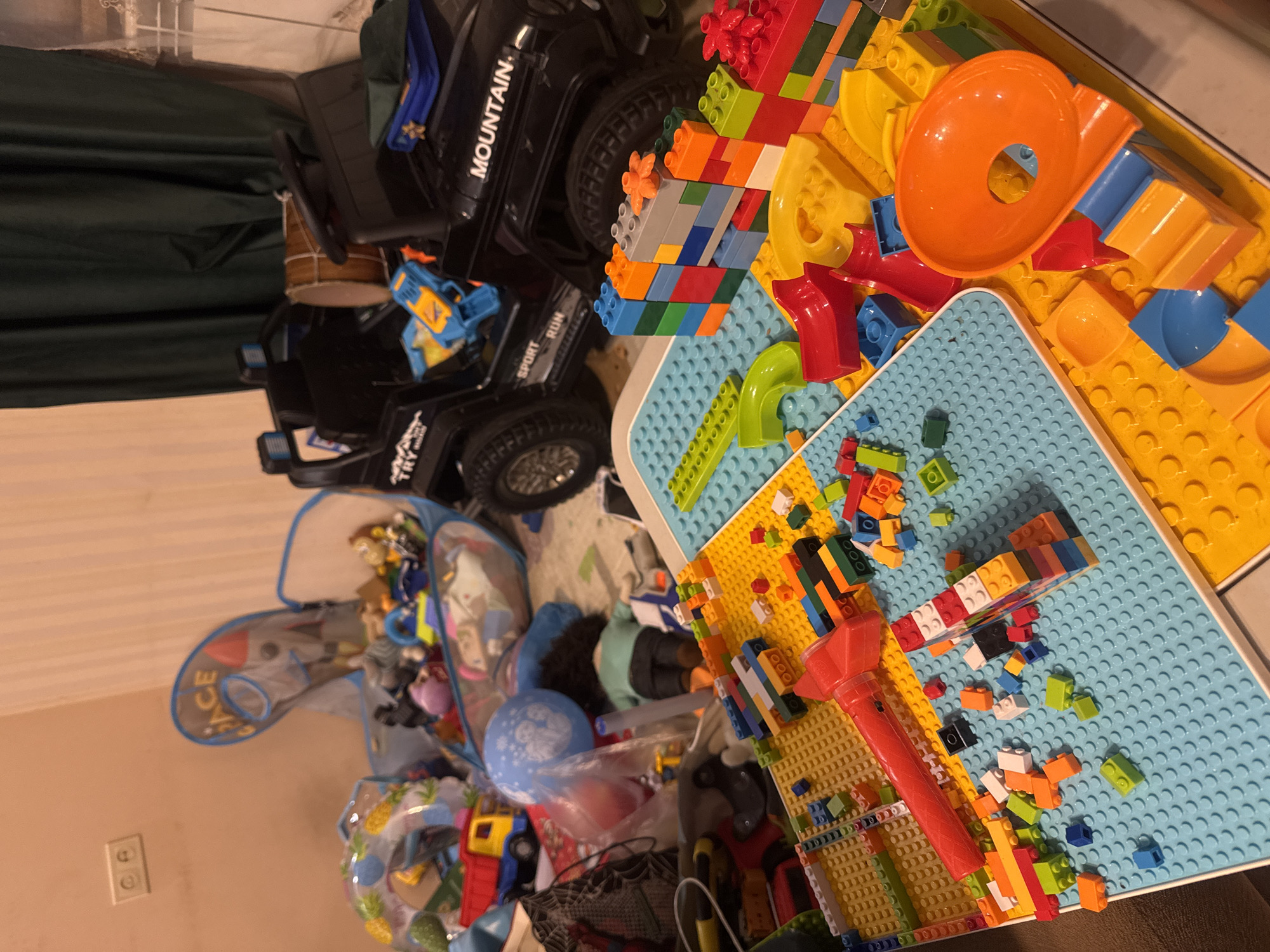The image shows a room in disarray. A black toy jeep with "MOUNTAIN" written on the side sits next to a colorful assortment of Lego blocks. A blue and yellow marble run is partially assembled on a blue and yellow baseplate. A large mesh toy storage bag overflows with various toys, including a deflated blue balloon. The location appears to be a child's playroom indoors, presumably in Yerevan, Armenia, given the user's request. No people are visible.