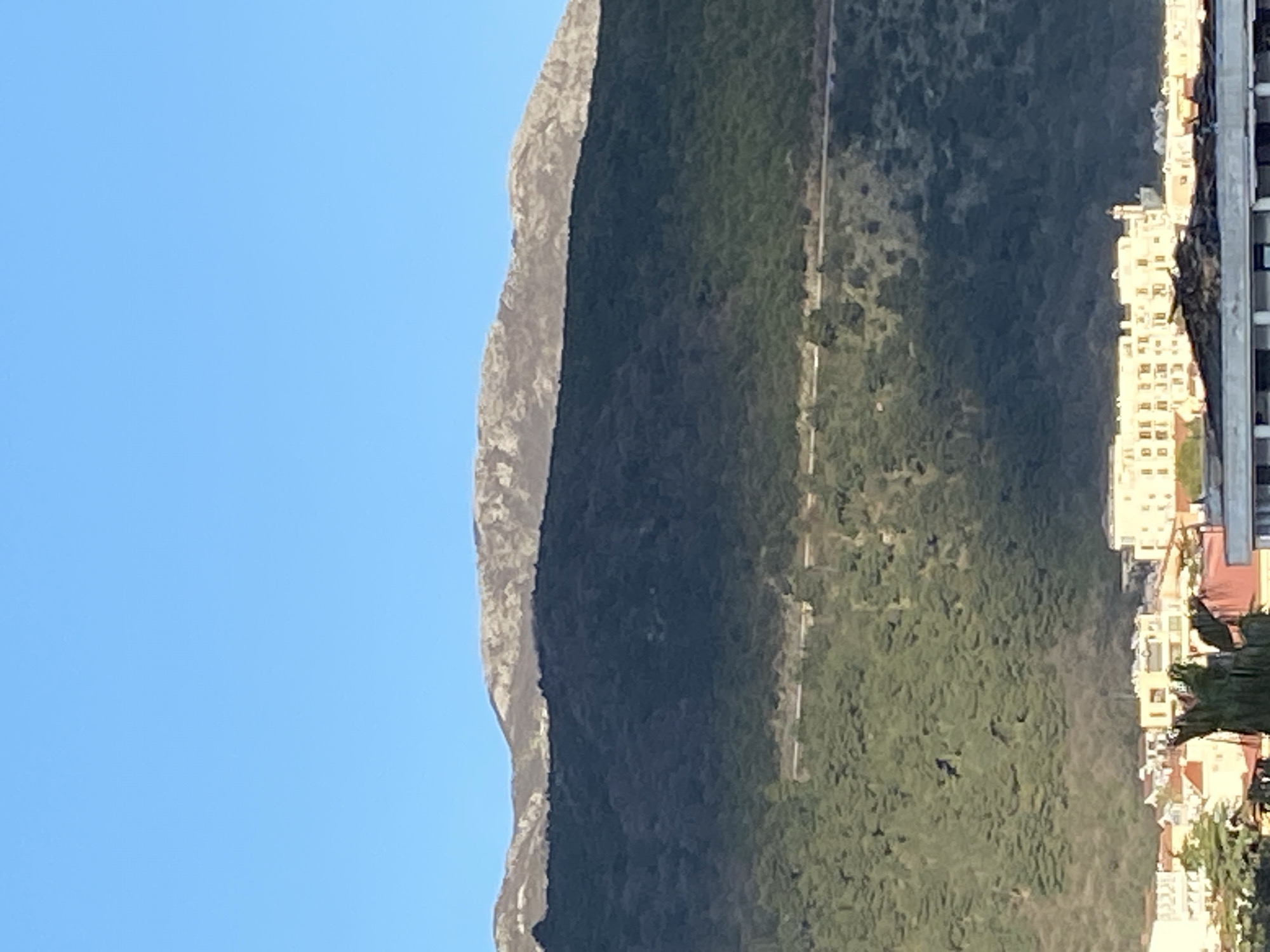 A vertical, medium shot captures a mountain range casting a shadow over a densely vegetated area and buildings in Budva, Montenegro, against a clear blue sky. The left side of the mountain range is sunlit, revealing its rocky texture, while the right side is in shadow, merging with the lush greenery below. A road or pathway is faintly visible along the mountain's slope. To the right, buildings with light-colored facades and red rooftops are arranged, some partially obscured by trees. In the foreground, the edges of a building or structure with a metallic element are slightly visible. The overall impression is a blend of natural landscape and urban development.