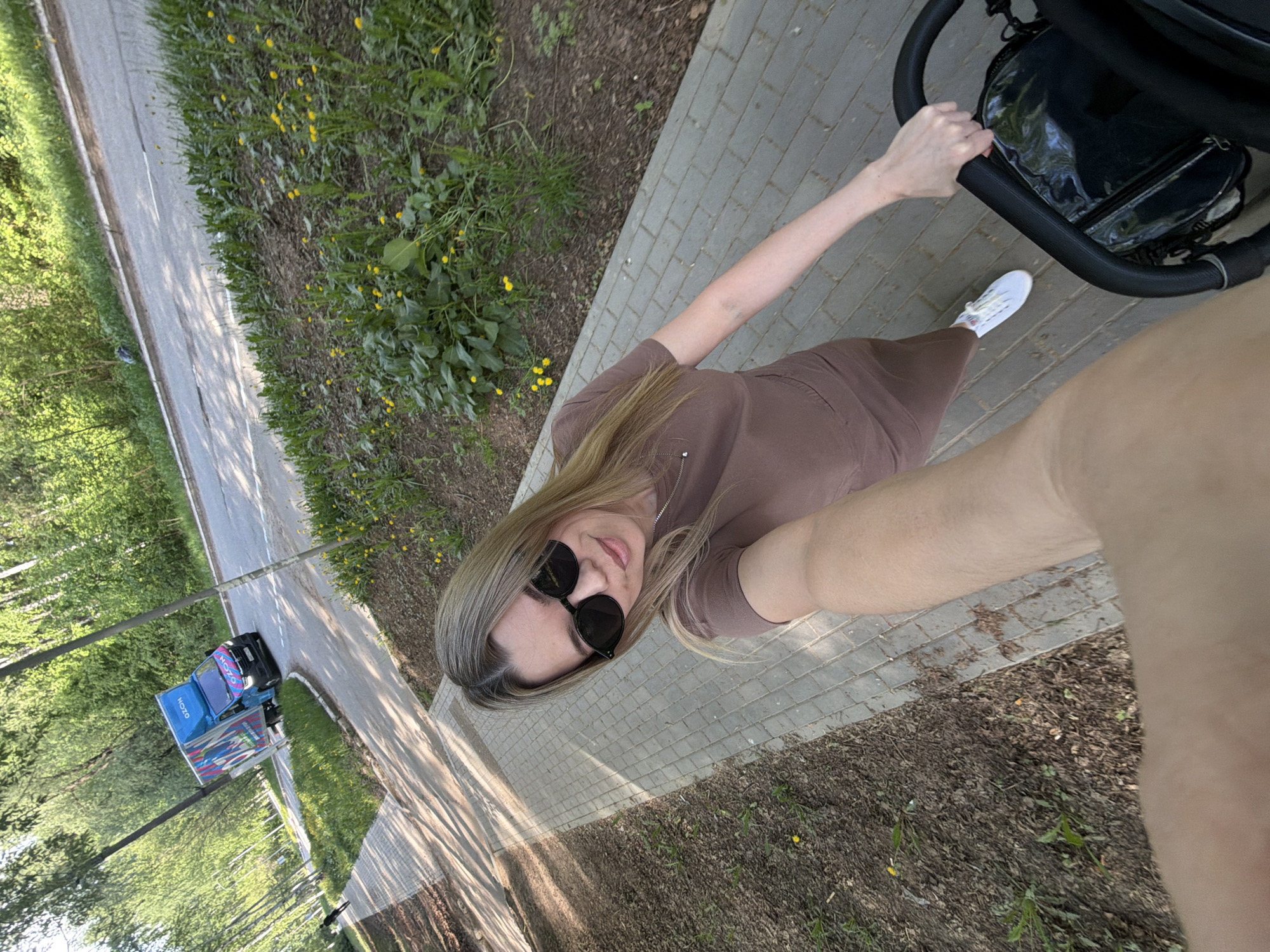 A woman wearing sunglasses and a brown dress is walking along a paved path while pushing a black stroller. The location is Chaikovsky, Russia. The path is lined with grass and wildflowers on one side, and trees and a road with a delivery truck on the other. The woman is holding her arm out to take a selfie.