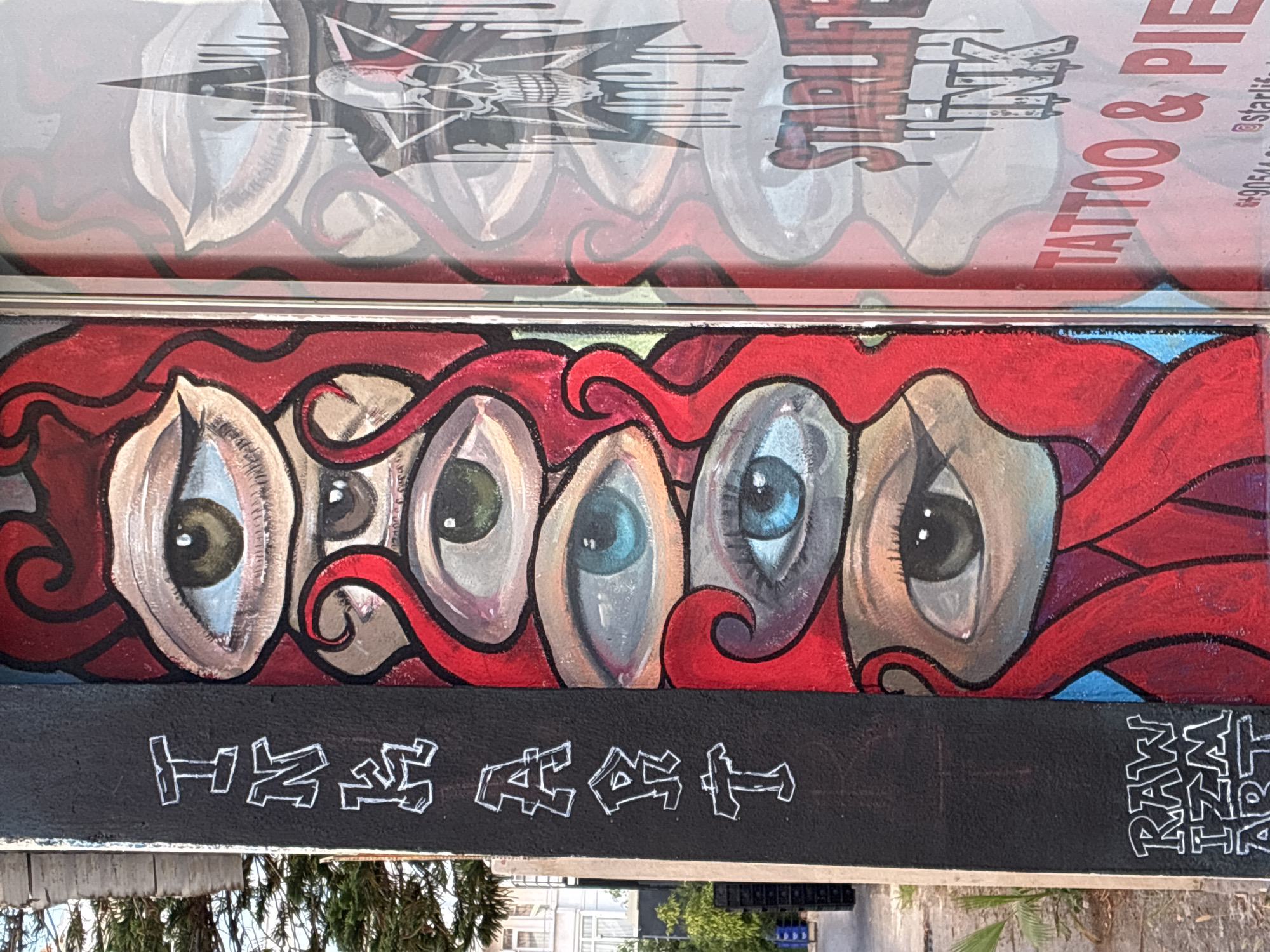 The image shows a mural on the side of a building in Alanya, Turkey. The mural features a series of stylized eyes with red, wavy hair. On the left side of the mural, the words "INK ART" are written vertically in white block letters. Below this, also in white block letters, are the words "RAW IZM ART". The mural is painted on a dark, textured surface. To the right of the mural, a window reflects an image of a skull with dripping ink, the words "STARLIFE INK TATTOO & PIERCE" and a phone number. The environment appears to be an urban street setting with a portion of a tree visible on the left.