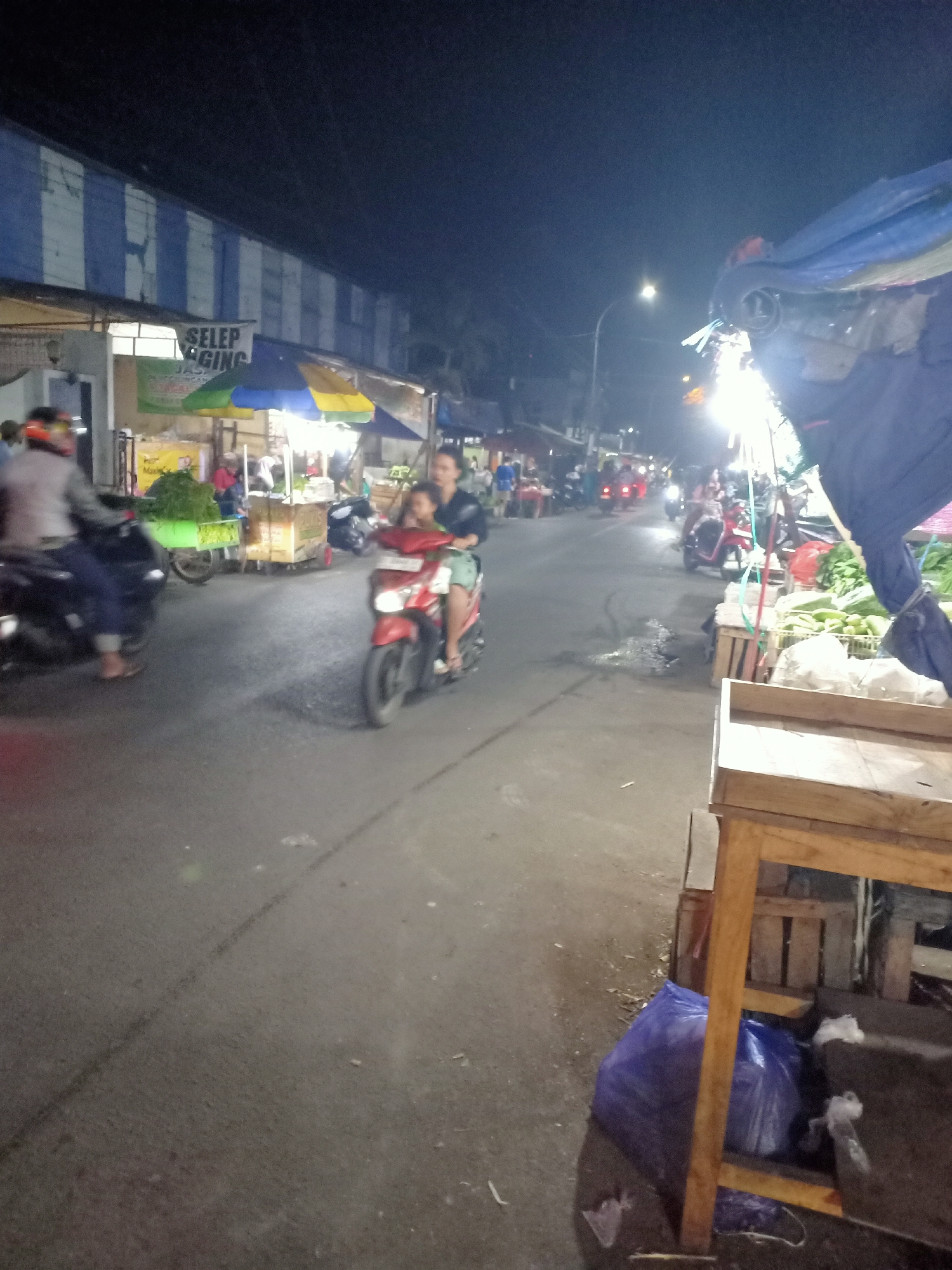 The image depicts a street scene at night in what is identified as Semanding, Indonesia. The foreground is dominated by a wooden stand or stall, partially obscured by a blue plastic bag and other debris on the ground. In the mid-ground, several motorcycles are present. One motorcycle, a red scooter, is moving away from the viewer with two occupants: a woman driving and a child seated behind her. Another motorcycle, dark in color, is parked or moving in the background to the left, with a person on it wearing a helmet.
The background shows buildings with a distinctive blue and white striped facade, suggesting a market or commercial area. Brightly lit stalls with umbrellas are visible, indicating a vibrant night market atmosphere. Various goods, including vegetables, are displayed on the stalls, with a large pile of green vegetables being particularly prominent. Streetlights and artificial lighting from the stalls illuminate the scene, casting a night-time ambiance. The road surface appears to be asphalt, with some wet patches and tire marks visible. The overall impression is one of a bustling, active night market in a local Indonesian setting.