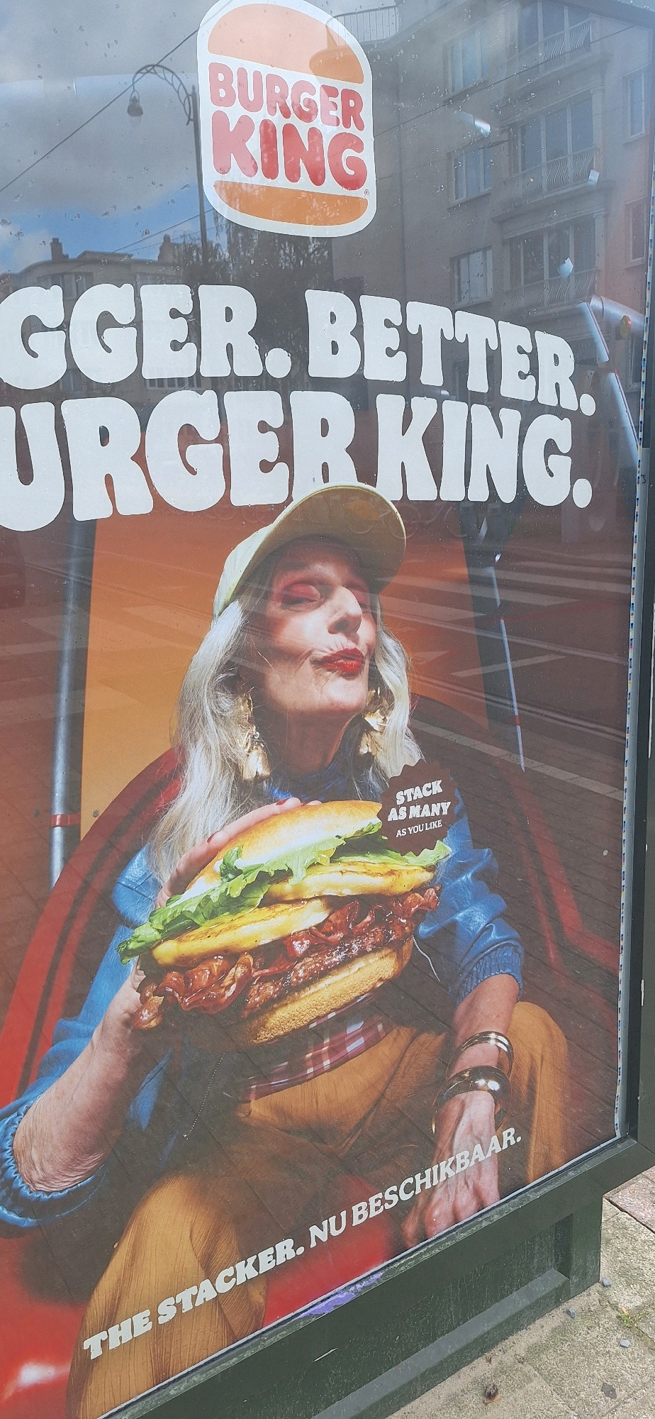 The image shows a Burger King advertisement poster displayed in Ixelles - Elsene, Belgium. The poster features a close-up of a person with blonde hair, wearing a yellow cap and a blue jacket, holding a large burger. The person has dramatic makeup with red lipstick and eyeshadow. Text on the poster includes "BURGER KING," "GGER. BETTER. URGER KING.", "STACK AS MANY AS YOU LIKE," and "THE STACKER. NU BESCHIKBAAR." The background of the poster shows a blurred urban environment with buildings and some kind of red structure. The poster is mounted on a dark green frame and is exposed to the elements, as indicated by water droplets on the surface. The ground in front of the poster is a pavement with some debris.