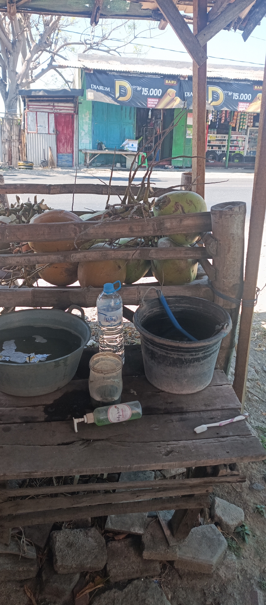 The image shows an outdoor scene under a simple wooden shelter, likely in Jatisari, Indonesia. In the foreground, a wooden platform holds several objects. To the left, a large, dark grey basin is filled with water. Next to it sits a glass jar, and to the right of that, a bottle of green liquid, possibly hand soap. Further to the right is a large, dark bucket containing water and a blue tube. On the far right of the platform, a toothbrush with pink and white bristles lies horizontally. Behind the platform, a display of coconuts is arranged on a bamboo structure. Some coconuts are brown, while others are green. Beyond the coconuts, a street is visible with a row of small shops. One shop has a red door, and others have colorful facades, including a large banner advertising "DJARUM RP 15.000". The overall environment appears to be a roadside stall or small shop, typical of a local commercial area in Indonesia. The lighting suggests it is daytime, with bright sunlight casting shadows.