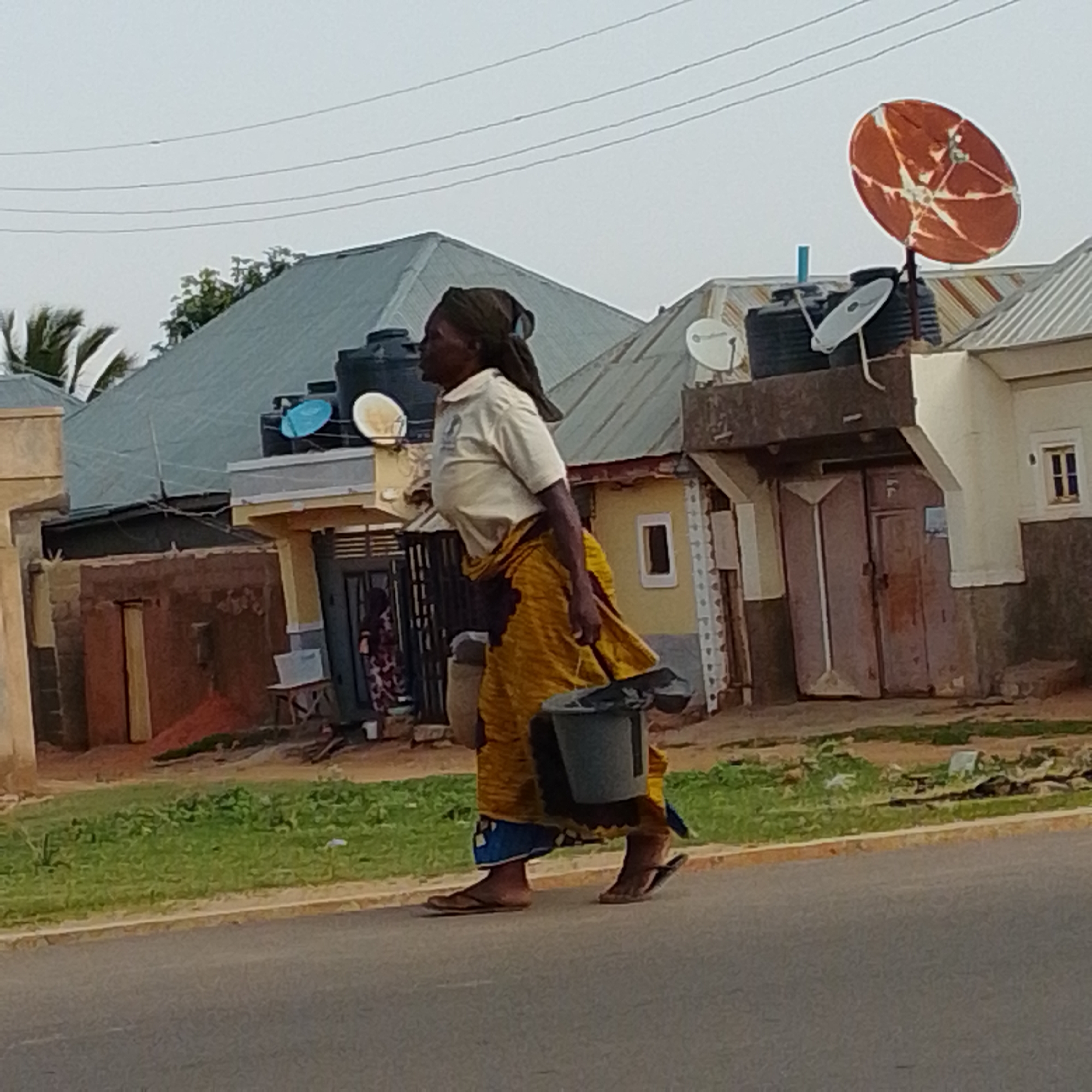 A woman walks along a paved road in the city of Gombe, Nigeria. She is wearing a light-colored shirt, a yellow patterned skirt, and a headscarf. She is carrying a gray bucket in her right hand, and her left hand is visible near her side. The woman is walking from left to right in the frame. The background shows several buildings with corrugated metal roofs, some with satellite dishes. There is a patch of green grass between the road and the buildings. The sky is overcast.