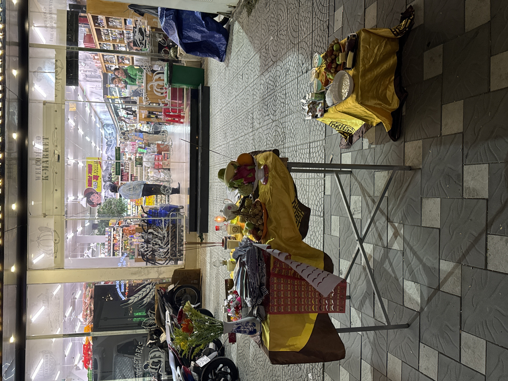 This is a street-level shot at night in front of a K-Market store in Da Nang, Vietnam. A metal frame table is set up on the sidewalk, covered with a yellow and brown cloth. On the table, there are offerings including a vase of flowers, incense sticks, a teapot, bowls of food, and a plate of fruit. To the right, another smaller table is covered in a yellow cloth with more offerings of fruit and small bowls. In the background, the brightly lit K-Market store has glass windows displaying shelves of products and customers inside. Several motorbikes are parked to the left of the frame.