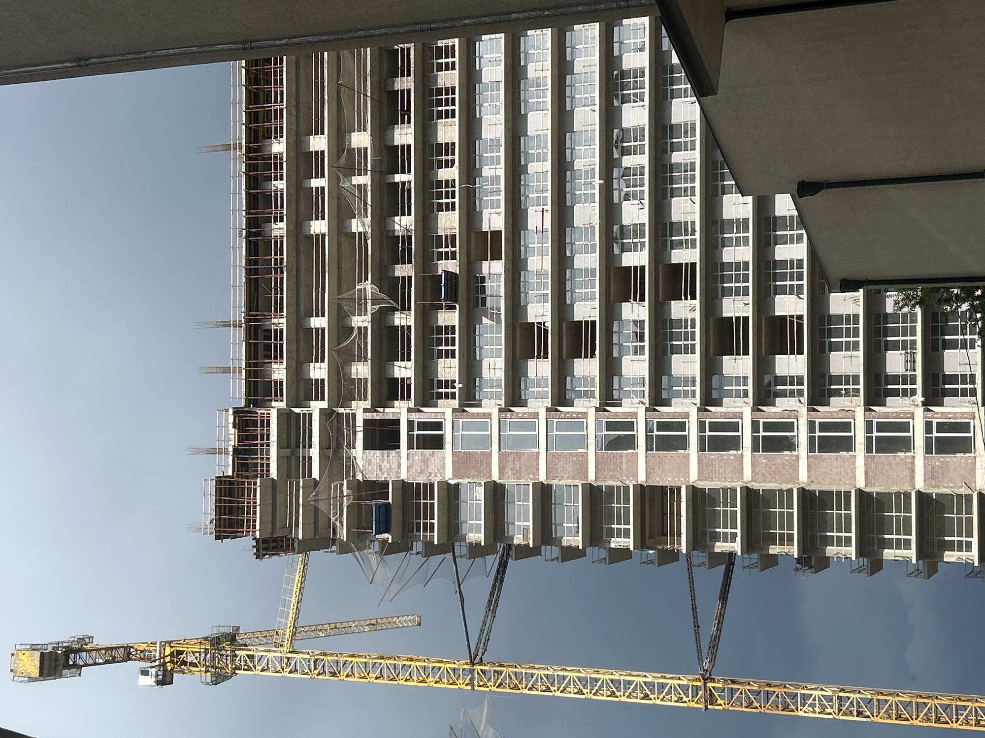 The image shows a tall building under construction in Tbilisi, Georgia. The building's facade is characterized by a grid of rectangular window openings. Scaffolding is visible on one side of the building. A yellow crane dominates the foreground. The sky above ranges from light blue to a darker overcast tone. The viewpoint is from below, looking up at the building and crane.