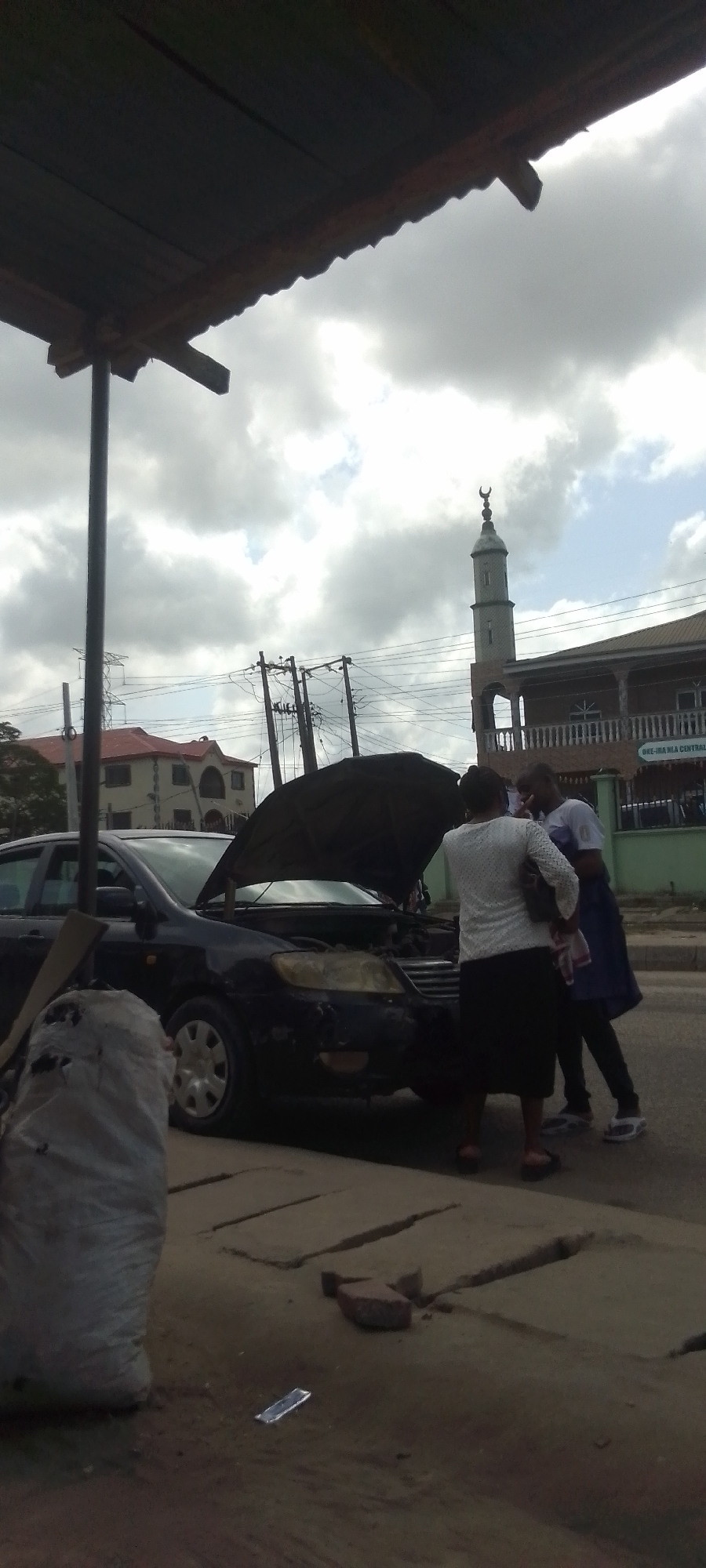 The image depicts a daytime scene with a black car parked on the side of a street. The hood of the car is open, and two people are standing near the engine. One person is wearing a white top and a black skirt, while the other is wearing a white shirt and a blue overall. In the background, there is a building with a minaret, suggesting a mosque, and another building with a red roof. There are utility poles and power lines visible in the background as well. A bag of material and a brick are on the ground near the car. The image is taken from under a shelter of some sort. The location appears to be a city in Unknown, Unknown. The sky is overcast with grey and white clouds.