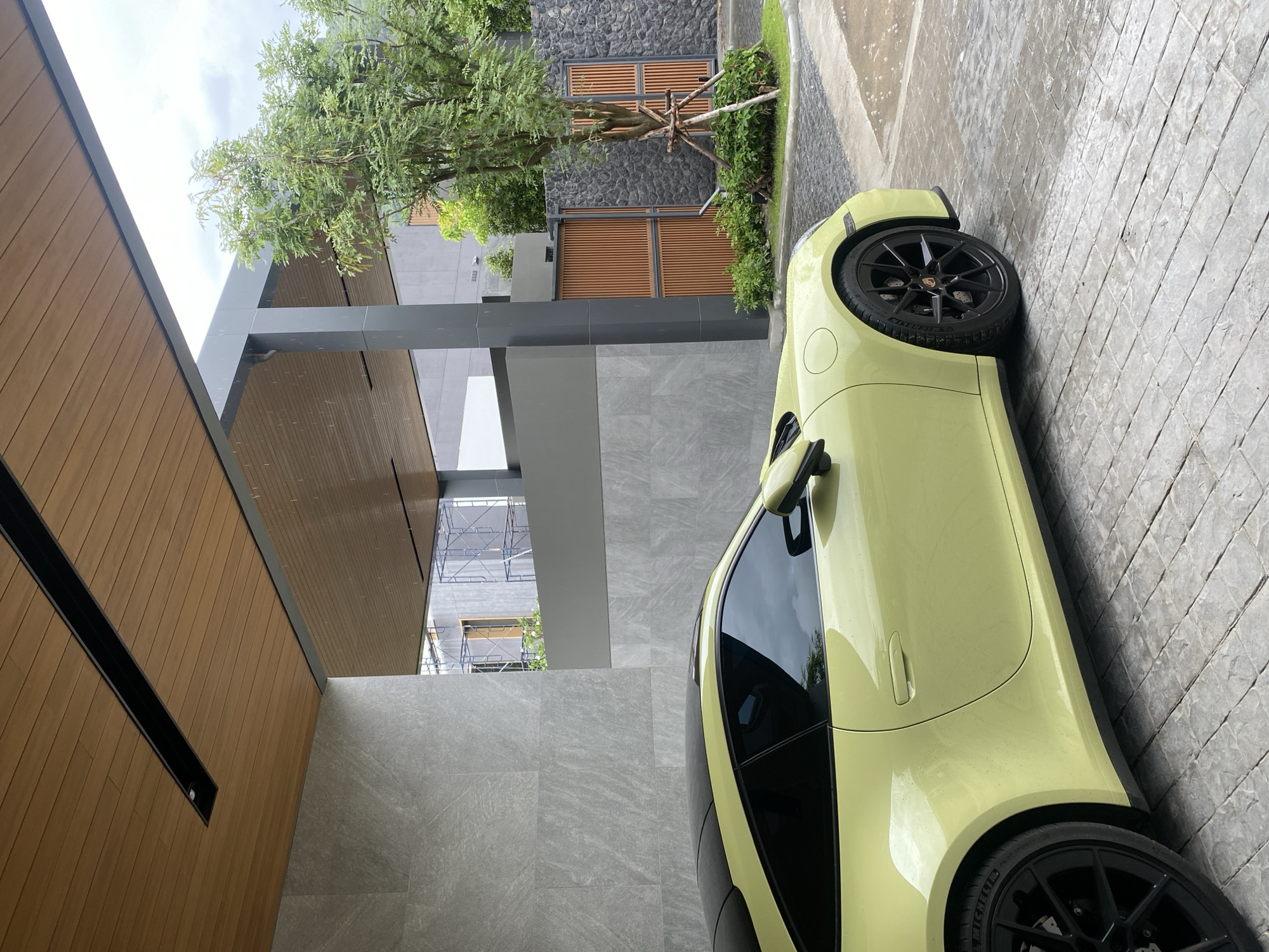The image shows a yellow Porsche car parked in front of a modern building with gray and wood paneling in Choeng Thale, Thailand. The ground is paved with gray stone tiles. A small tree and some greenery are visible next to the building. Scaffolding is visible in the background. The sky is cloudy.