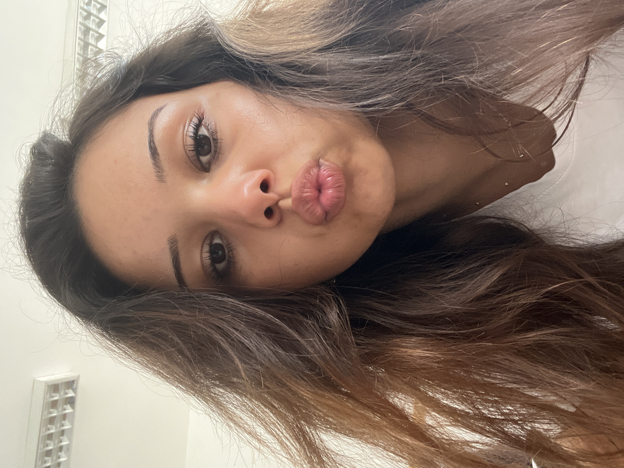 A close-up portrait shows a person, likely female, making a "kiss face" or "duck face" with pursed lips directed towards the camera. The image is tilted, with the subject's head leaning to the left side of the frame. Her brown eyes are open and gaze directly at the viewer, highlighted by visible eyelashes and well-groomed, dark eyebrows. Her complexion appears smooth.
Her long, wavy hair cascades around her, featuring a color gradient from dark brown roots to lighter brown and blonde tones at the ends, suggesting an ombre or balayage style. The hair appears somewhat tousled. In the background, a plain white wall is visible, along with a portion of a modern, rectangular ceiling light fixture with a grid design. The lighting is bright and evenly illuminates her face. There are no clear visible accessories or text in the image. The overall mood conveyed by the expression is playful and direct.