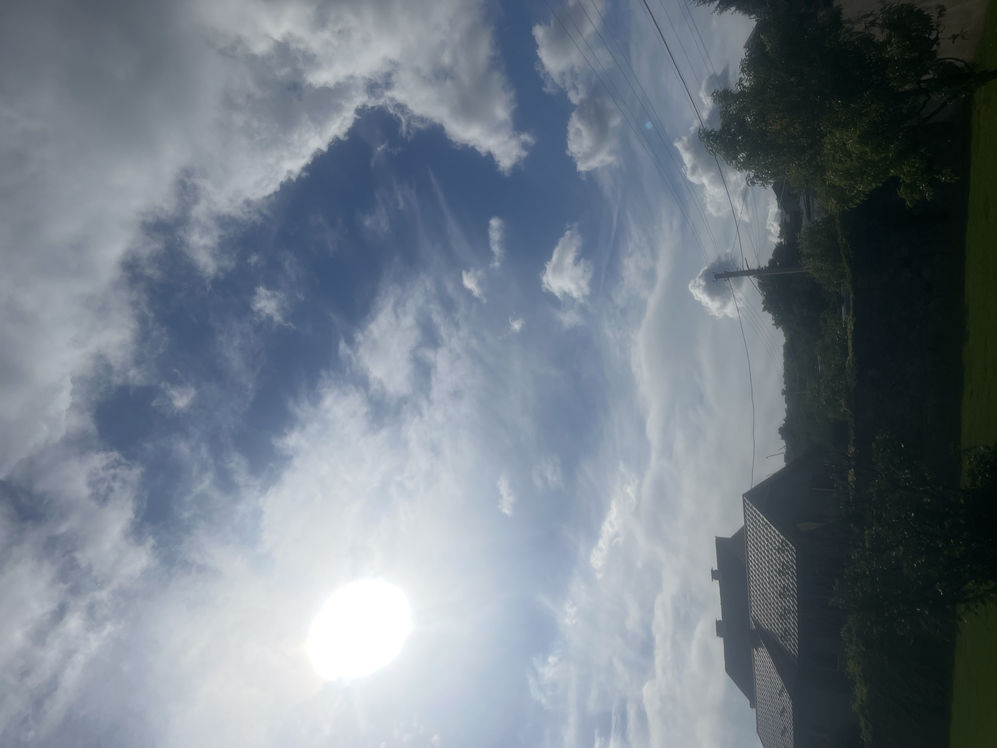 The image captures a partially overcast sky over a rural or suburban landscape, likely Onokivtsi, Ukraine, given the provided context. The sun is visible as a bright, diffused disc in the upper left quadrant, casting a bright light. Wispy clouds are scattered across the blue sky, with larger, more defined clouds present in the upper right.
In the lower half of the image, silhouetted against the sky, are the roofs of several houses with dark-tiled roofs and chimneys. These structures appear to be residential. Dense green foliage, including trees and bushes, forms a dark band in front of the houses and extends into the foreground, obscuring the ground level. A few utility poles with wires are visible in the middle ground. The overall impression is a daytime scene with a mix of sun and clouds. There are no people or animals visible in the image.
