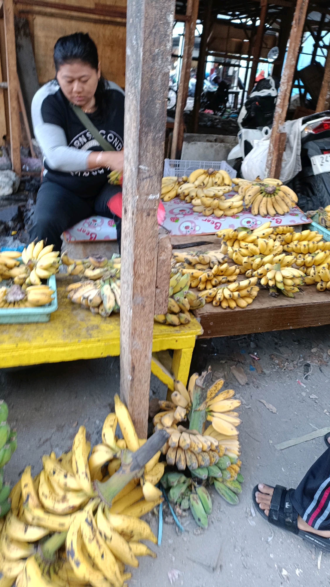 A woman is selling bananas at an outdoor market in Surabaya, Indonesia. She is wearing a black shirt with gray sleeves and dark pants, and she is sitting on a low stool. She is holding a bunch of bananas, and there are many more bunches of bananas displayed on tables in front of her. The market appears to be somewhat cluttered, with wooden structures and various goods for sale. In the background, motorcycles and other vendors can be seen. The ground is dirt and debris.