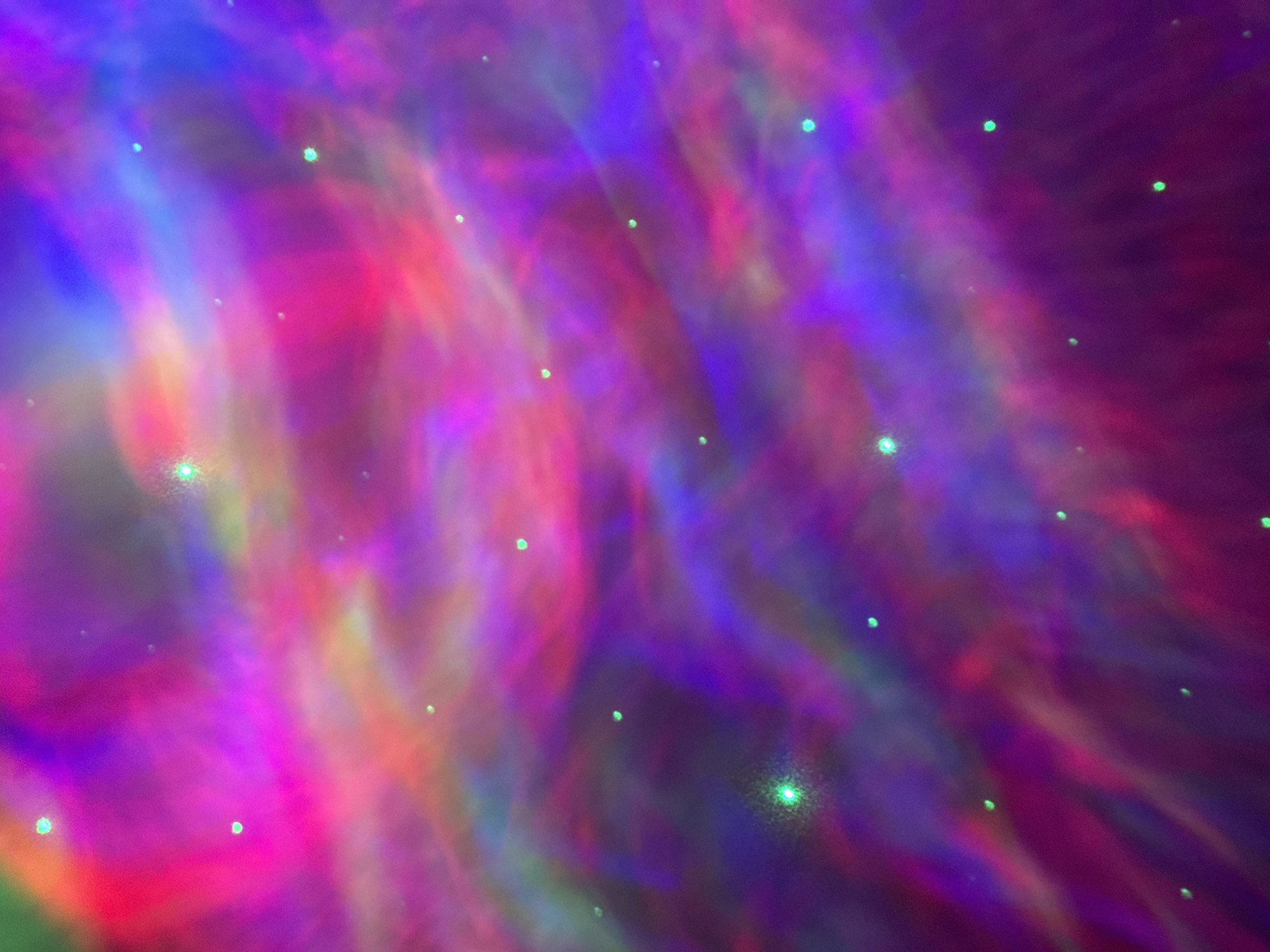 The image shows a projection of colorful lights, predominantly pink, purple, and blue, resembling a nebula or aurora borealis. Scattered throughout the projection are small, bright green dots that appear to be stars. The lighting is dynamic and appears to be in motion, creating swirling patterns. The overall impression is abstract and otherworldly. There is no indication of the city of Tourcoing, France, or any specific location within it. The image focuses solely on the light projection itself.