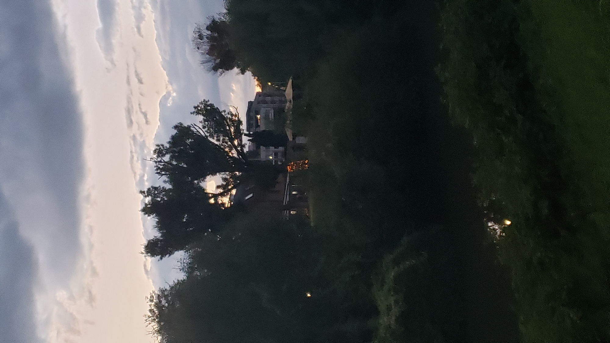 The image depicts a twilight scene viewed through foliage. In the background, a residential building with illuminated windows is visible, situated behind a cluster of trees. The sky is overcast with dramatic clouds. There is a faint reflection on a body of water in the foreground, possibly a river or pond. The overall impression is a somewhat obscured view of an urban setting at dusk, with natural elements encroaching on the man-made structures. The location is specified as Prešov, Slovakia.
