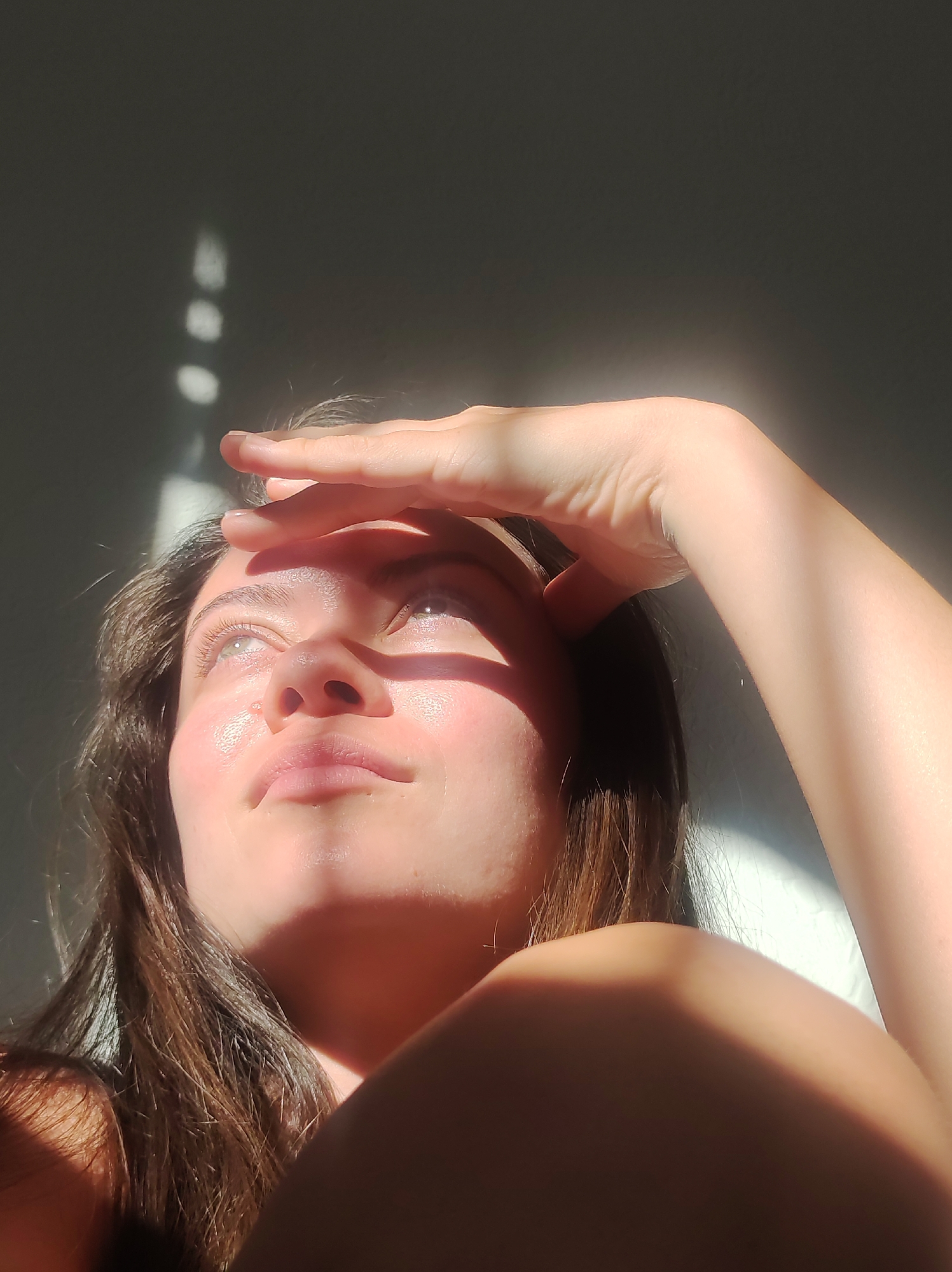 The image shows a close-up of a young woman's face, tilted upwards, with her right hand shielding her eyes from a bright light source. Sunlight casts dramatic shadows across her face and the background, creating streaks of light and dark on a plain, gray wall. Her skin appears dewy, reflecting the light. Her eyes are open and looking towards the light. Her brown hair frames her face. The overall impression is one of a candid moment captured in natural light, possibly indoors. The image does not contain any discernible elements that would indicate a specific location within Chaikovsky, Russia.