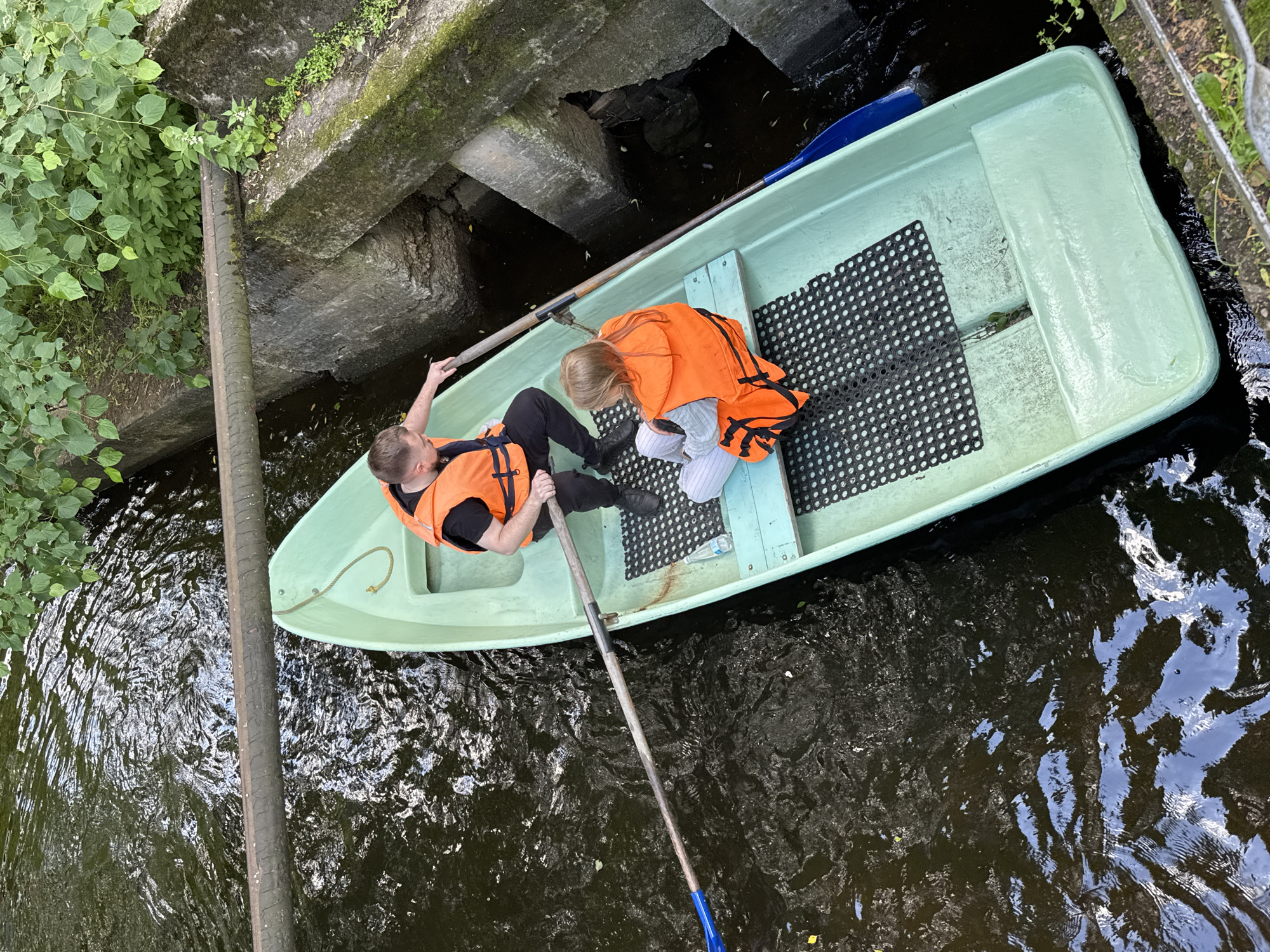 Two people, a man and a girl, are in a light green rowboat on a narrow body of water, likely a canal or river in Saint Petersburg, Russia, as indicated by the architectural style of the surrounding stone structures and lush greenery. The man, positioned at the rear of the boat, is holding a wooden oar with a blue tip, actively rowing. He is wearing an orange life vest over a dark t-shirt and black pants, with short, light brown hair. The girl, seated in the front of the boat, also wears an orange life vest and is dressed in striped pants and a light-colored top. She is looking towards the stern of the boat. The water is dark and appears somewhat murky, with ripples and reflections. On either side of the waterway, there are substantial concrete or stone embankments, overgrown with vegetation, including trees and bushes. A thick, weathered pipe runs horizontally across the scene, suspended above the water. The lighting suggests it is daytime, with natural light filtering through the foliage.