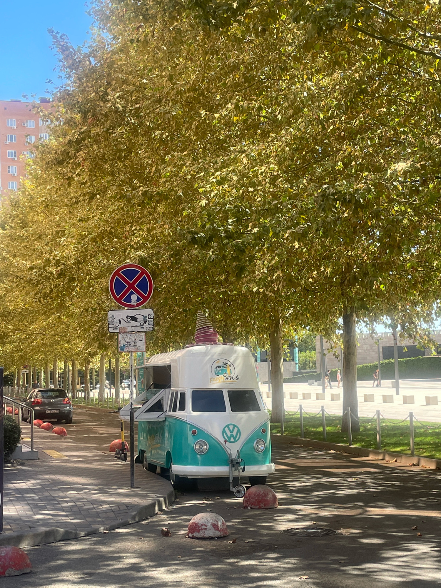 This is a medium-shot, eye-level, outdoor photograph of a street scene in Krasnodar, Russia, on a sunny day. The foreground features a sidewalk and a street. On the sidewalk, a turquoise and white Volkswagen Type 2 van, repurposed as a food truck, is parked. It has a conical hat on its roof and a logo on the front that reads "FRESH MOBILE". Several red, dome-shaped bollards are scattered on the street and sidewalk.
Behind the food truck, a row of trees with golden-yellow leaves lines the street, creating a canopy. A traffic sign with a red circle and a blue cross, indicating "no stopping", is visible above the sidewalk. Further down the street, a dark-colored car is parked on the left.
In the background, there are modern buildings, sidewalks, streetlights, and a few people walking. The overall impression is a pleasant, urban setting with a touch of autumn scenery. The photograph was likely taken on a clear day, given the bright sunlight and blue sky.
