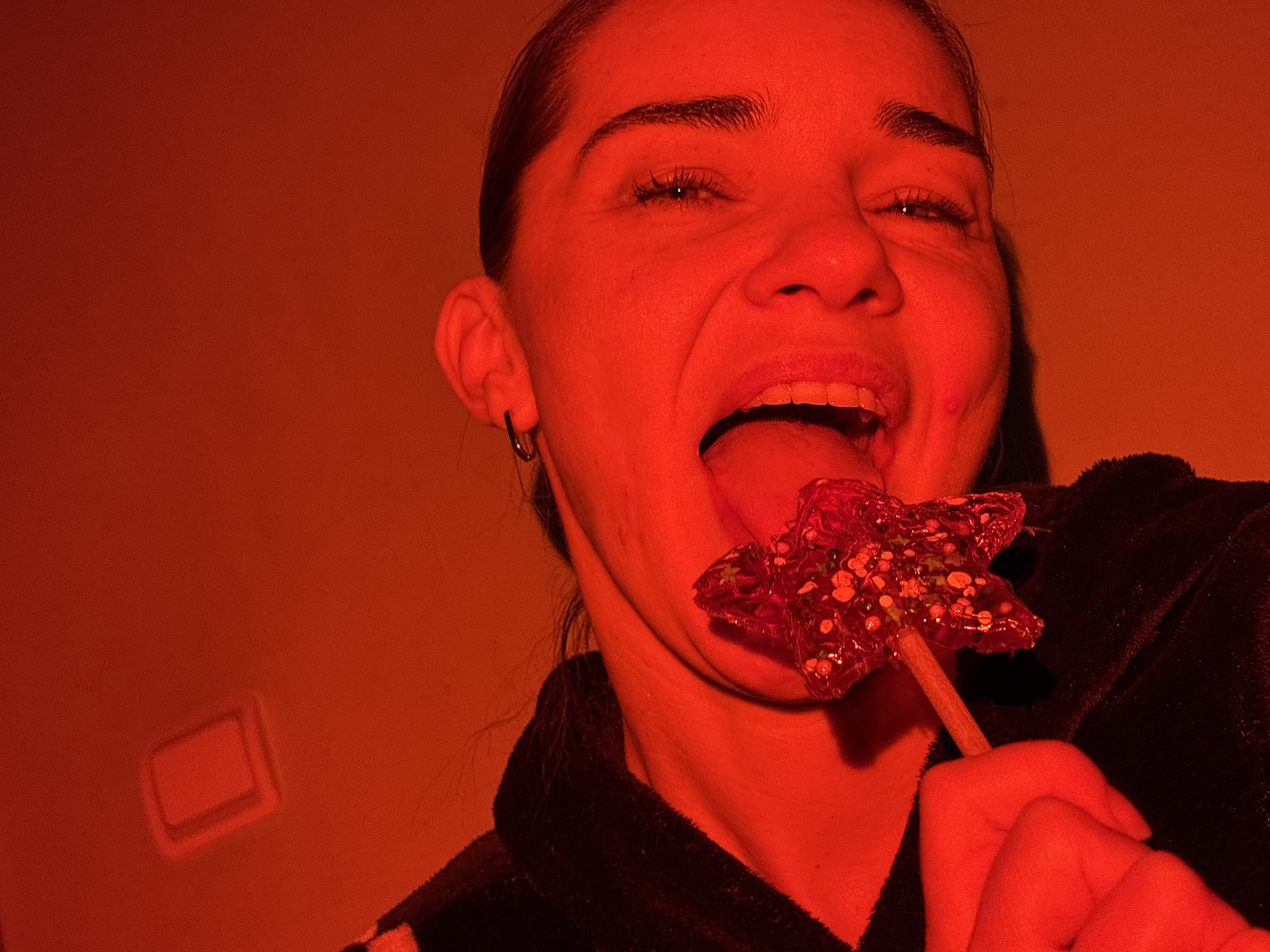 A person is depicted in a close-up shot under a predominantly red lighting condition. The individual is holding a star-shaped lollipop and is in the act of licking it with their tongue extended. The person is wearing a dark-colored, possibly velvet robe. The background appears to be a plain wall with a rectangular light switch or electrical outlet visible. The location is unknown.