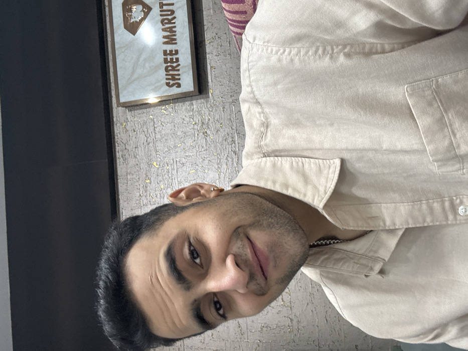 A man faces the camera with a slight smile. He has dark hair, brown eyes, and light stubble. He wears a light beige button-up shirt, with a silver chain visible at his neck. Behind him is a textured wall with a sign reading "SHREE MARUTI" in gold lettering. A dark-colored object is partially visible on the left edge.