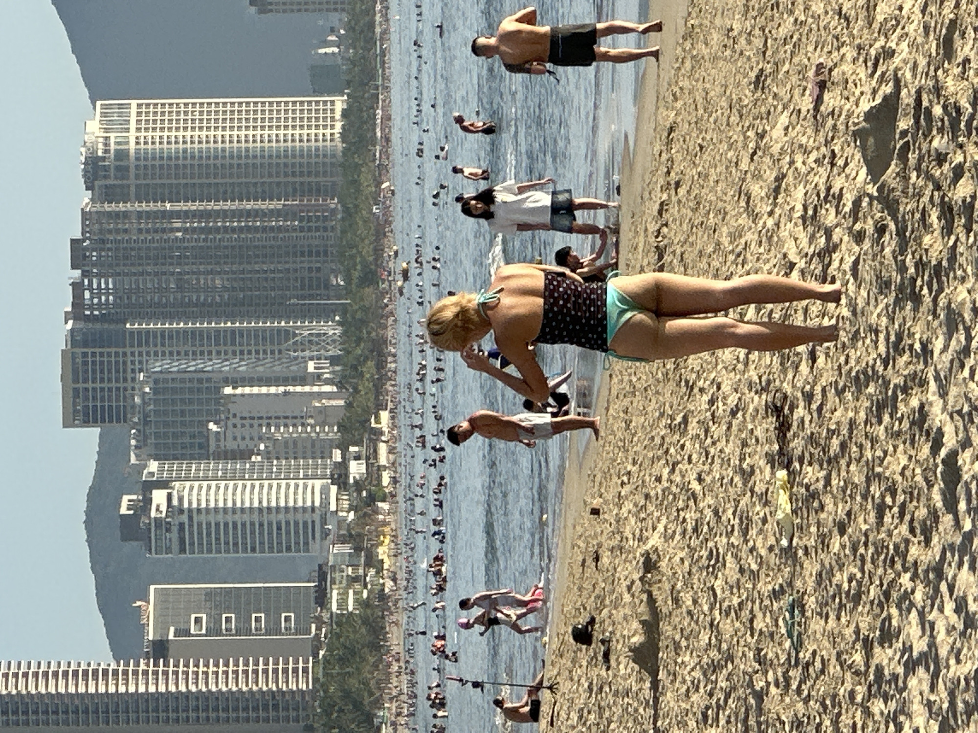 This image captures a bustling beach scene in Đà Nẵng, Vietnam, with the city's modern skyline forming a backdrop. In the foreground, a woman in a teal bikini is standing on the sand, facing away from the camera, with her hand near her hair. The beach is populated with numerous people enjoying the water and sand. Some are swimming or wading in the ocean, while others are walking along the shore or sitting on the beach. The water appears calm, and there is a gentle surf. In the background, several tall, contemporary buildings rise above a line of trees, indicating an urban setting. The sky is clear and blue, suggesting a sunny day. The overall impression is one of a lively and popular tourist destination.