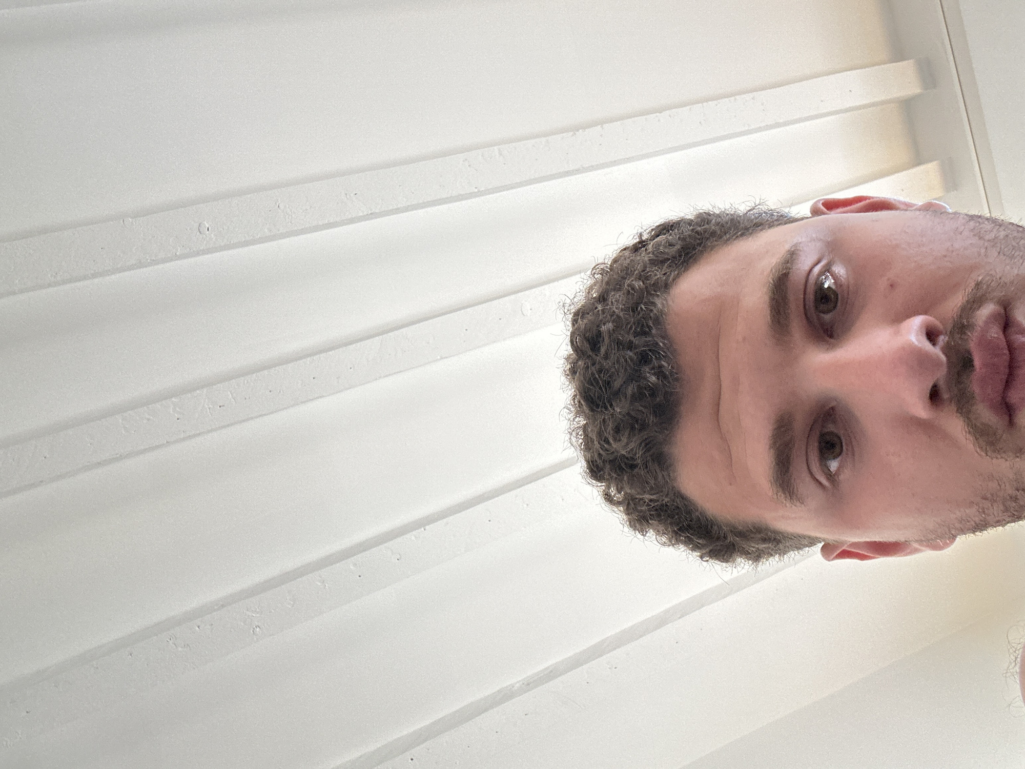 A close-up, angled shot shows a person's face, partially visible in the lower-right corner, with curly hair and stubble, looking up at the camera. The background consists of a white, paneled surface, likely a ceiling or wall, with horizontal lines and subtle textures. The environment appears to be indoors. There is no evidence to suggest that the location is in Milan, Italy.
