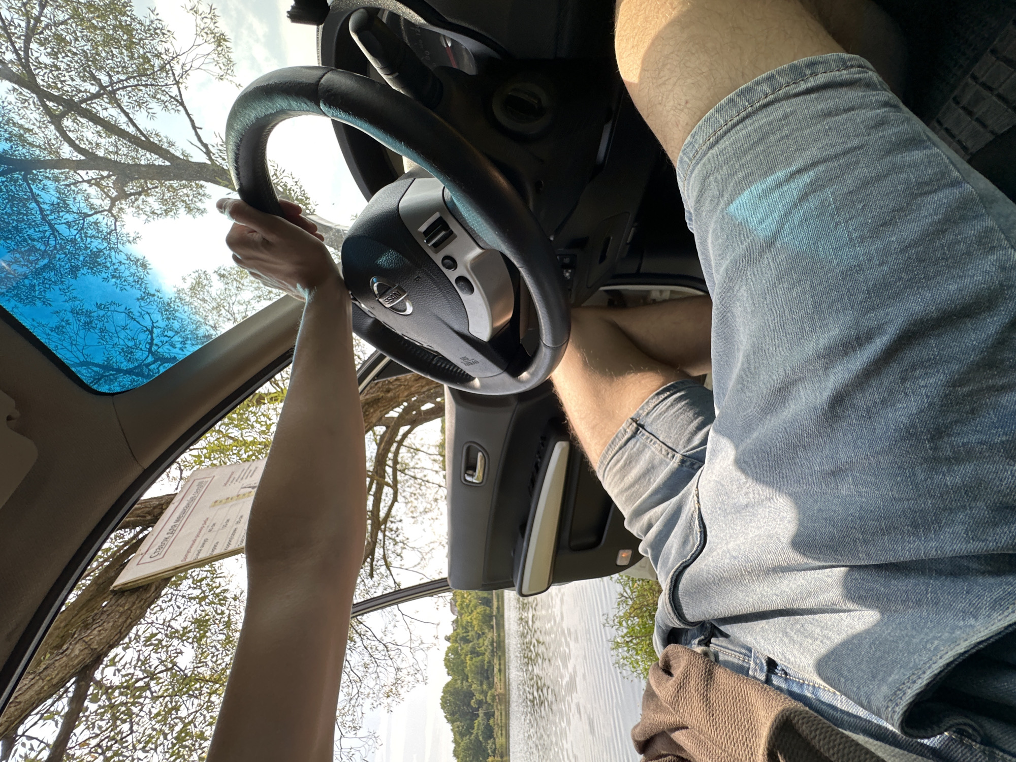 An outdoor, eye-level shot taken from inside a vehicle, shows a person's hands on the steering wheel and their legs in denim shorts. The open car door reveals a lake and trees in the background. The scene appears to be near Makiv, Ukraine, given the context of the request.