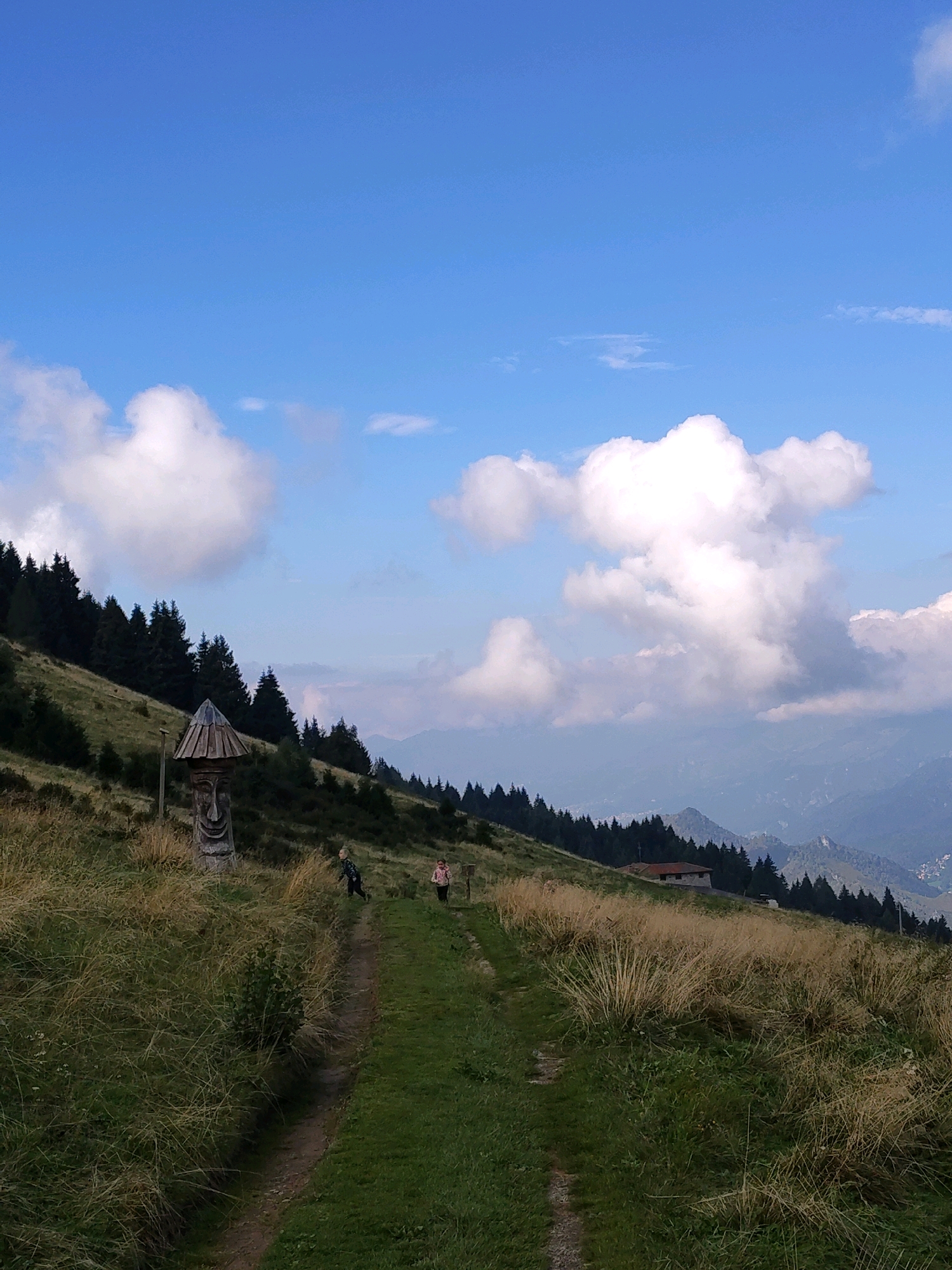 A sunny day with scattered clouds is depicted in this landscape image. The scene is set on a grassy hillside, with a path winding through the vegetation. In the foreground, a carved wooden figure stands near the path. Two individuals, a child and an adult, are visible walking along the path further up the hill. In the background, a cluster of evergreen trees lines the hillside, and beyond them, distant mountains are visible under a clear blue sky. A small building with a red roof is nestled among the trees in the middle ground.
