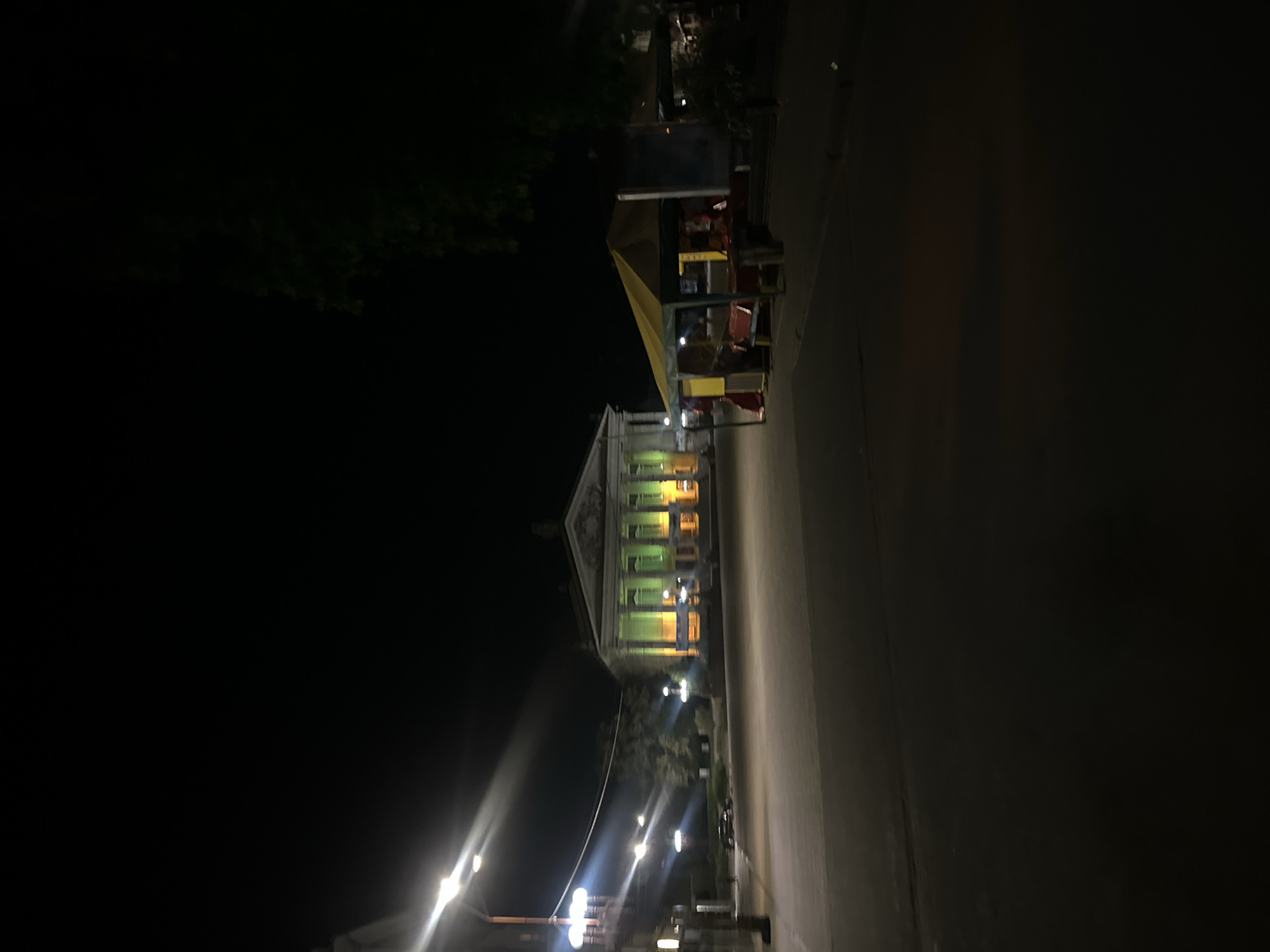 At night in Ternopil, Ukraine, an illuminated building with a classical facade stands beside a street. Streetlights line the road leading to the building. A kiosk or market stall with a yellow awning is positioned near the building. Trees are visible in the periphery. The setting is urban, with architectural and commercial elements apparent.