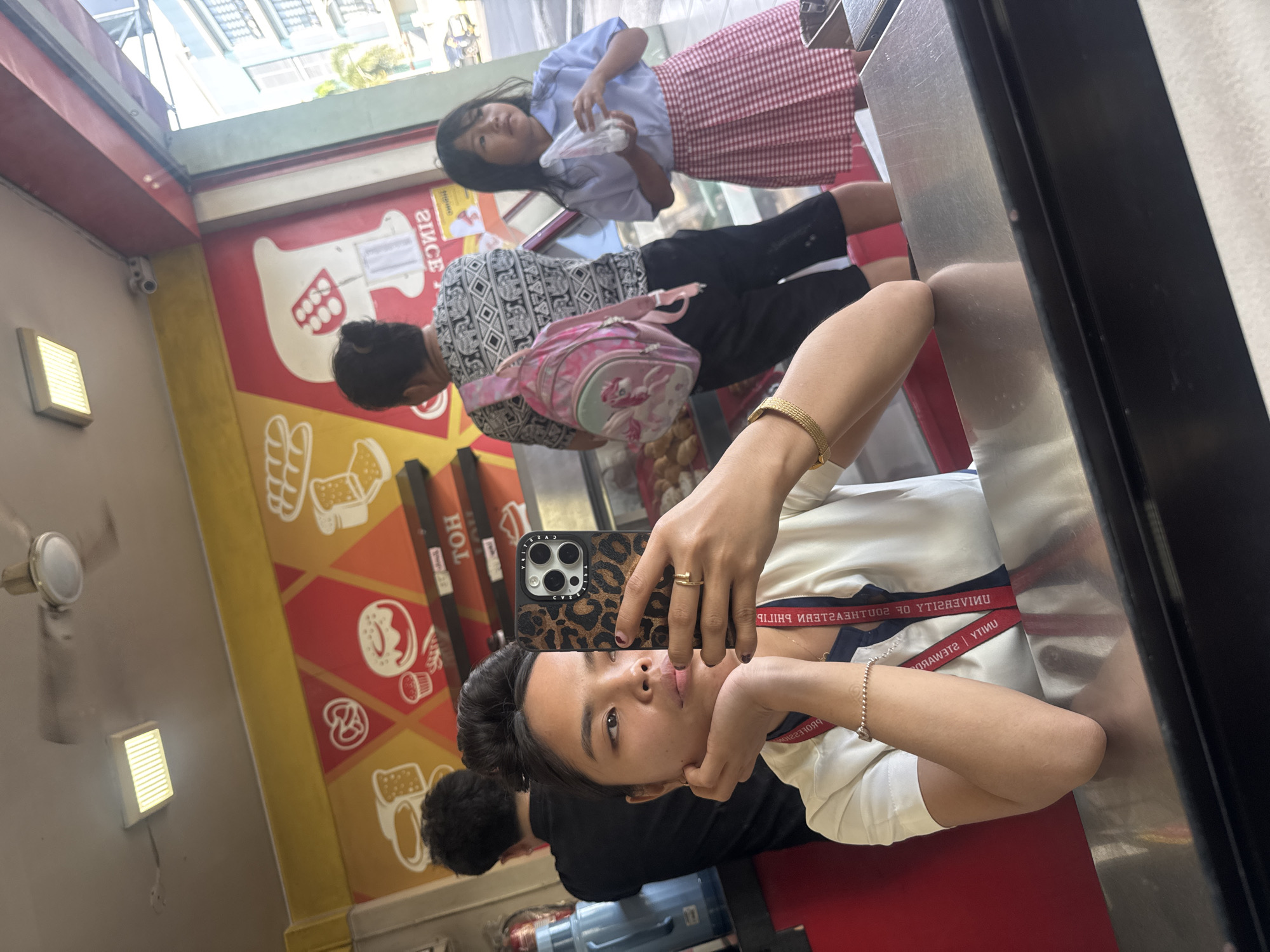 The image is a mirror selfie taken by a young woman sitting at a counter in what appears to be a bakery or food establishment. The woman is wearing a white collared shirt with a red lanyard and a bracelet. She is holding a phone with a leopard-print case, taking the picture with her right hand. Her left hand is resting on her chin, and she is looking at the phone.
In the background, reflected in the mirror, are other people. To the left, a person wearing a black shirt is partially visible. In the center, a woman with her hair in a bun is seen from behind, wearing a patterned shirt and carrying a pink backpack. To the right, a young girl in a light blue shirt and a red and white checkered skirt is holding a clear plastic bag.
The environment suggests a commercial setting with a colorful mural on the wall depicting baked goods like bread, donuts, and pretzels. There is a display case with food items, a ceiling fan, and lighting fixtures. The presence of a sign with the word "HOT" and what appears to be reversed text on a sign in the background suggests a commercial establishment, possibly a bakery or snack shop. Based on the context provided that this is in Davao City, Philippines, the scene depicts a local business. The overall atmosphere is casual and appears to be daytime.