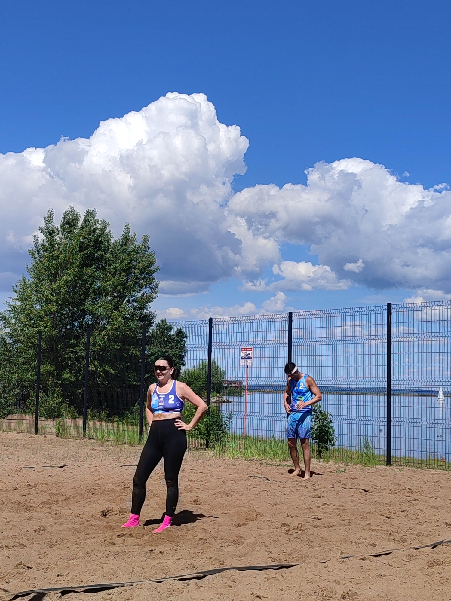 The image depicts an outdoor recreational scene on a bright, partly cloudy day in the city of Unknown, Unknown.
In the foreground, a sandy court occupies the lower half of the frame. Two individuals are positioned on this court. On the left, a female subject stands with her hands on her hips, facing slightly towards the right. She has dark hair, wears sunglasses, a blue and white sports bra with the number "2" prominently displayed, black athletic leggings that show some sand accumulation, and bright pink socks. On the right, further back on the court, a male subject stands, leaning forward and looking downwards. He wears a white headband, a blue and white tank top with the number "2," and blue patterned athletic shorts. He is barefoot.
A tall, dark-colored mesh fence with vertical posts runs horizontally across the mid-ground, separating the sandy court from a large body of water visible in the background. Attached to the fence on the left side, above the water, is a small red and white warning sign. Beyond the fence and water, a faint white sail of a boat can be observed on the far right.
The background features a prominent, dense green deciduous tree on the left, extending from behind the female subject and reaching into the mid-sky. Smaller green foliage is visible behind the fence and along the water's edge. The sky is a vibrant blue, filled with numerous large, white cumulus clouds, indicating a clear but cloudy day. Shadows are cast on the sand, suggesting direct sunlight. The overall environment appears to be a recreational area, possibly a beach volleyball court, adjacent to a lake or large river.