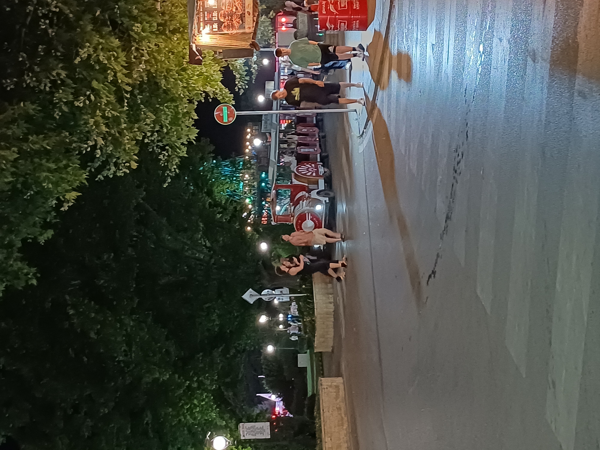 The image shows a nighttime scene in Varna, Bulgaria, likely on a pedestrian street or promenade. The foreground features a wet asphalt road with a pedestrian crossing. In the midground, a small, red and white train-like vehicle, possibly a tourist tram, is parked. Several people are visible walking and standing around the train and on the sidewalk. To the right, a food stand with a sign displaying prices and images of food items is illuminated. The background is dominated by lush green trees with some lights strung among them, suggesting an outdoor entertainment or dining area. The overall atmosphere is lively and suggests a tourist-oriented location.