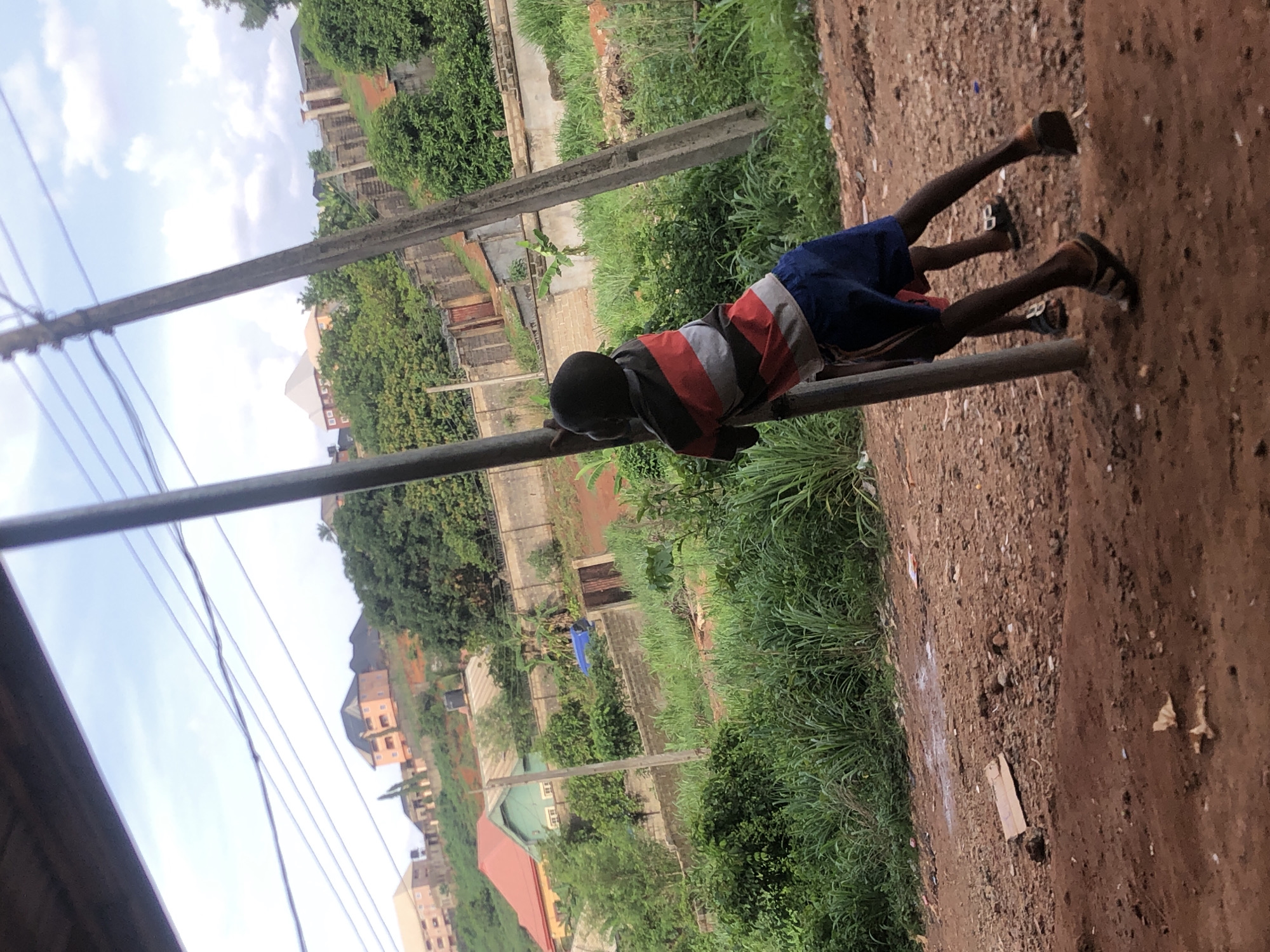 The image depicts a young boy balancing on a metal bar in Nkwelle Ezunaka, Nigeria. The boy is wearing a striped shirt and blue shorts, with sandals on his feet. He appears to be using his hands to grip the bar for support.
The background shows a residential area with houses, vegetation, and utility poles. The ground beside the bar is dirt, suggesting an unpaved area.