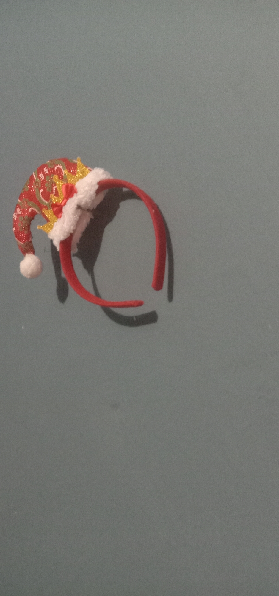 A Christmas-themed headband with a red hat, white trim, and gold accents is positioned on a gray wall. The hat is designed to resemble a Santa hat, complete with a white pom-pom at its tip and a small red bow. The headband itself is red. The image is cropped to focus on the headband, with the background showing a plain gray wall. The lighting casts a shadow of the headband onto the wall behind it. There are no people or other objects visible in the frame. The origin and specific location within Kendari, Indonesia, are not discernible from the visual information provided.