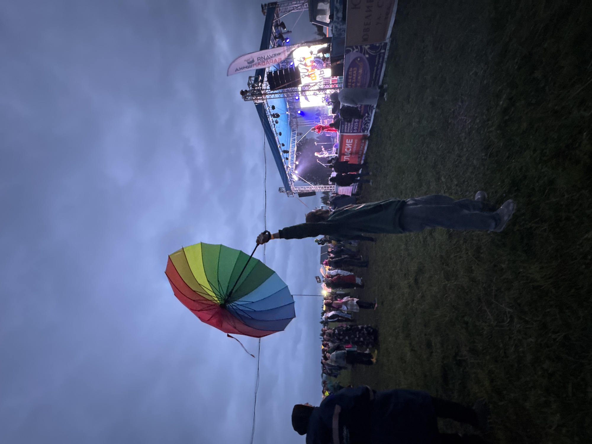 An outdoor music festival is underway in Bol'shaya Kivara, Russia, under an overcast, dark blue-grey evening sky. A person in the mid-right foreground, facing the stage, holds a large, open rainbow-colored umbrella aloft with their right arm. The umbrella displays distinct red, orange, yellow, green, blue, and violet panels. To the left, a person's back in a dark blue jacket is visible in the far-left foreground.
A crowd of attendees, dressed in casual attire, occupies the grassy field, largely facing a brightly lit stage on the right side of the frame. The stage features a blue canopy, metal trussing, and multiple spotlights illuminating a band performing. At least three musicians are discernible on stage, one prominently in a red outfit, with instruments such as guitars and drums visible. A large screen behind the performers displays the number "2024." Below the stage, a series of advertising banners with Cyrillic text, including "УЛАСИЕ" and "МЕБЕЛЬ НА ЗАКАЗ," are visible along a barrier. A white vertical flag with Cyrillic text stands to the right of the stage. A thin, dark cable stretches horizontally across the upper midground. The ground is a dimly lit grassy field.