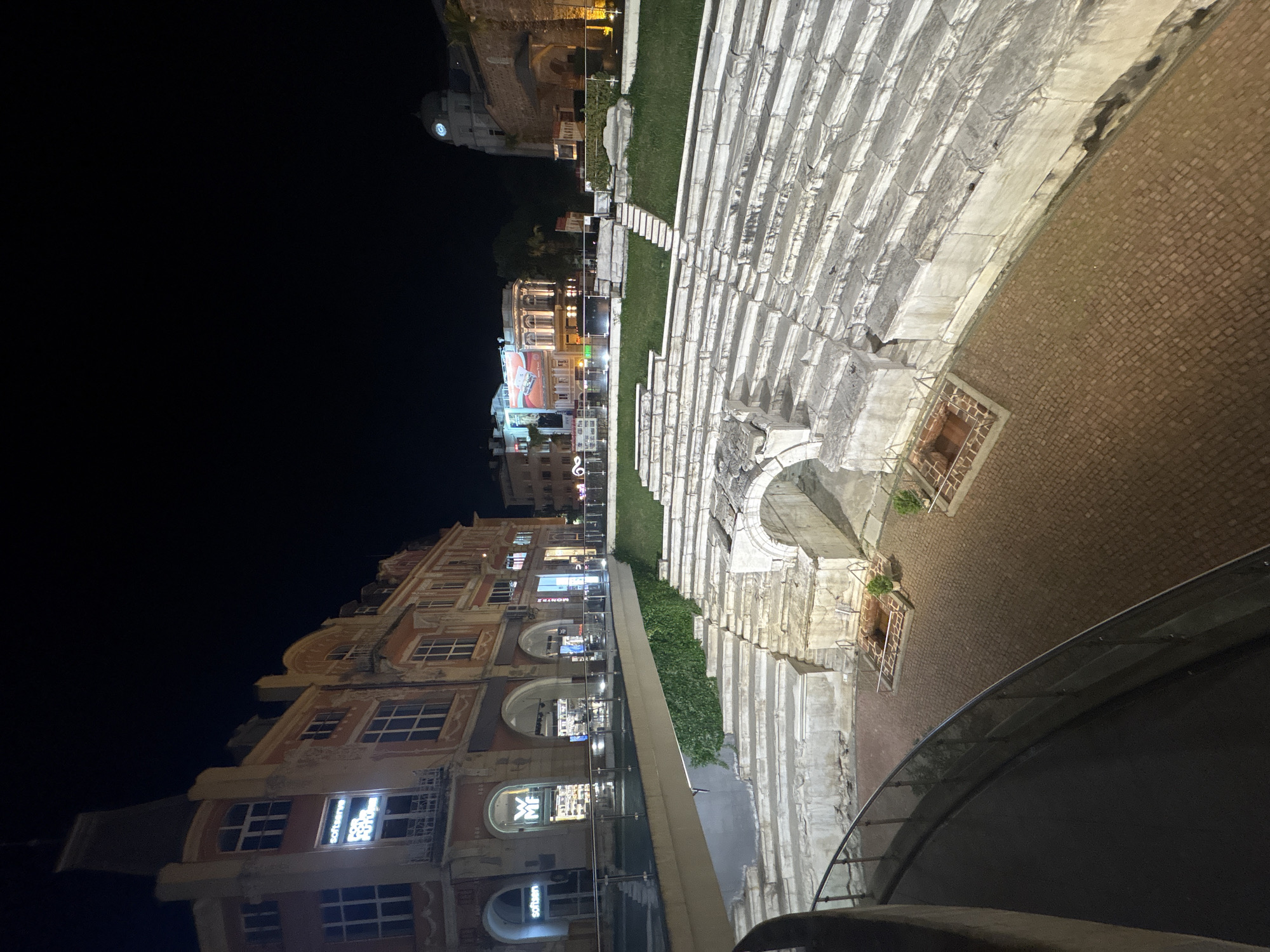 The image depicts the ancient Roman Theatre in Plovdiv, Bulgaria, at night. The stone seating tiers are visible on the right side of the frame, illuminated with a soft light. To the left of the theatre, a modern walkway with a glass railing is present. Beyond the walkway and seating tiers is a street lined with buildings. These buildings feature multiple stories and are illuminated with various lights from shops and businesses. The sky above the buildings is a dark expanse.